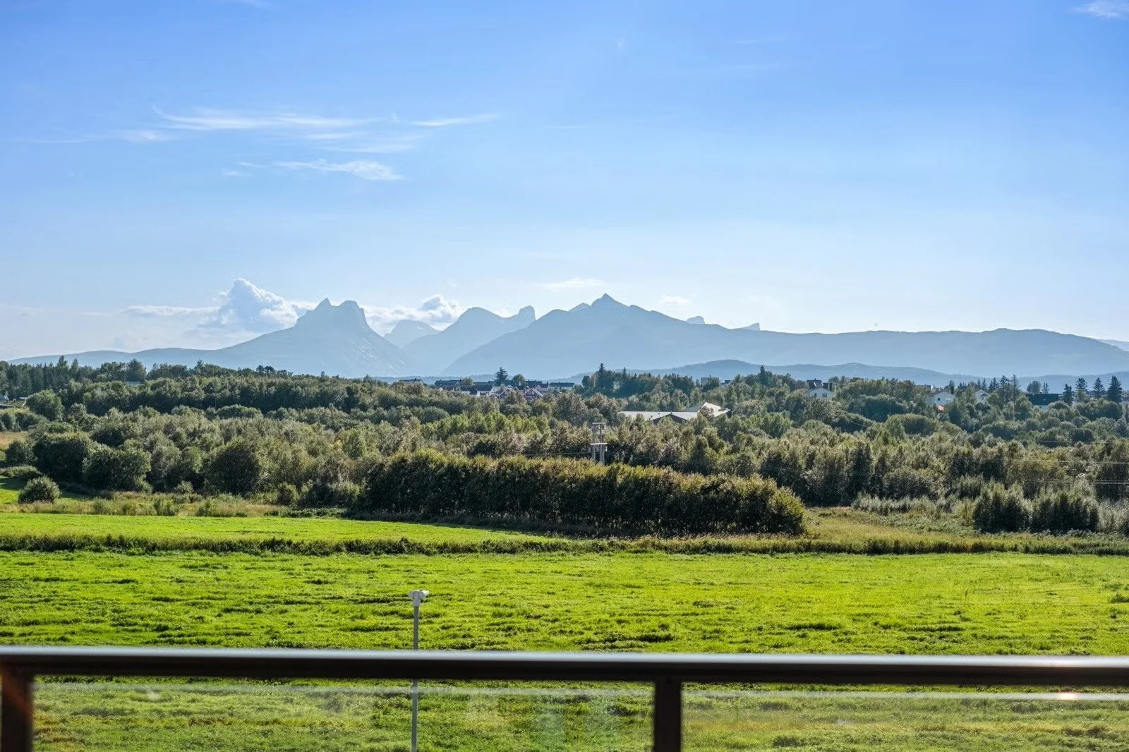 Scenic view of distant mountains with jagged peaks, green forest, and a clear blue sky with some clouds, seen over a lush green field.