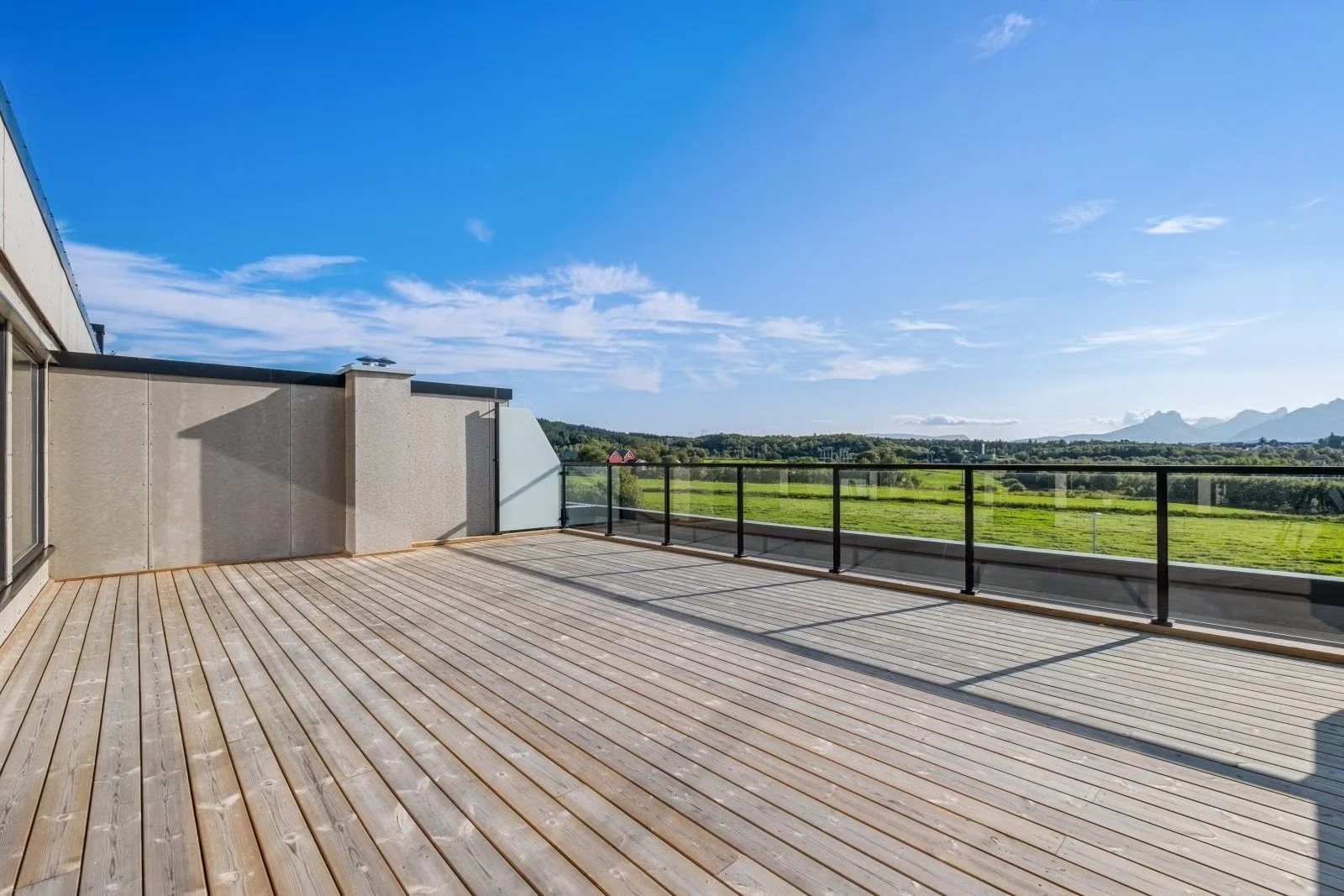 Empty balcony with wooden flooring, glass and metal railing, overlooking green fields and distant mountains under a partly cloudy blue sky.