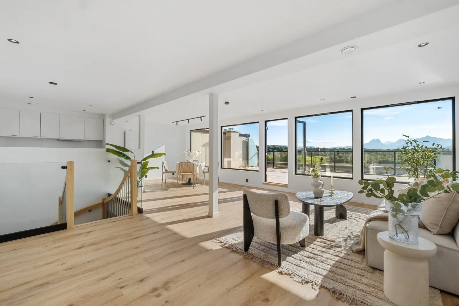 Bright, modern living room with large windows, wooden floors, beige and white furniture, and decor with plants and vases.