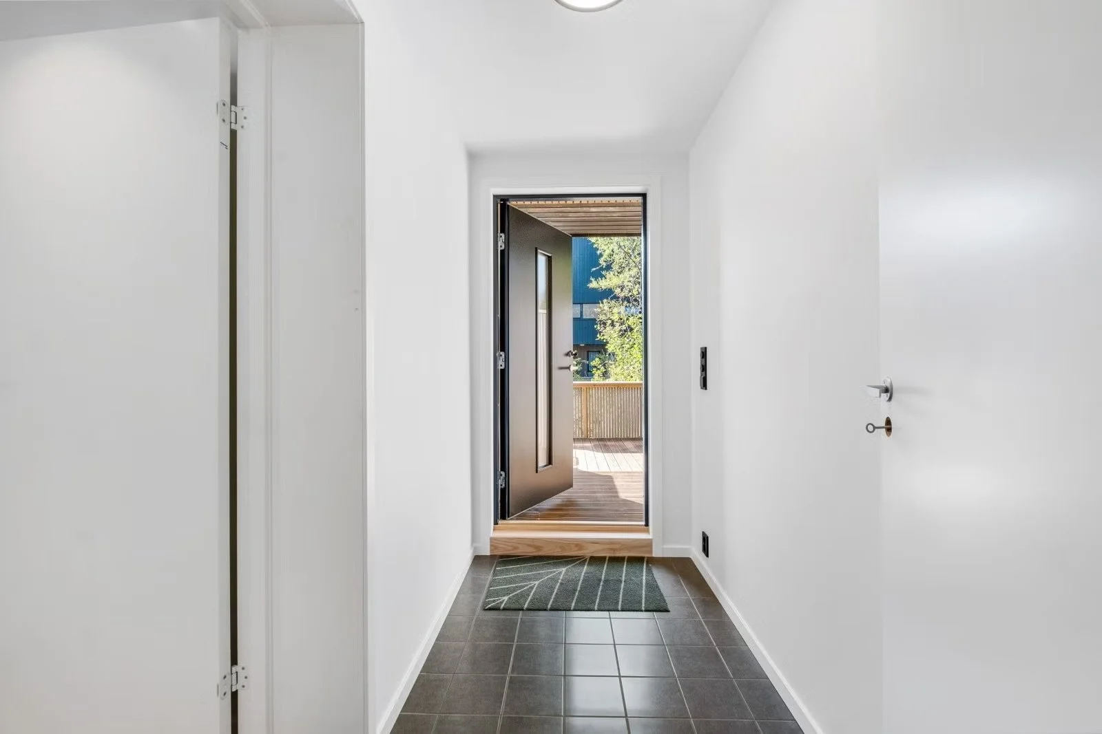 Interior view of a home entryway with white walls, a dark front door open to a porch, and a small green rug on dark tiled floor.