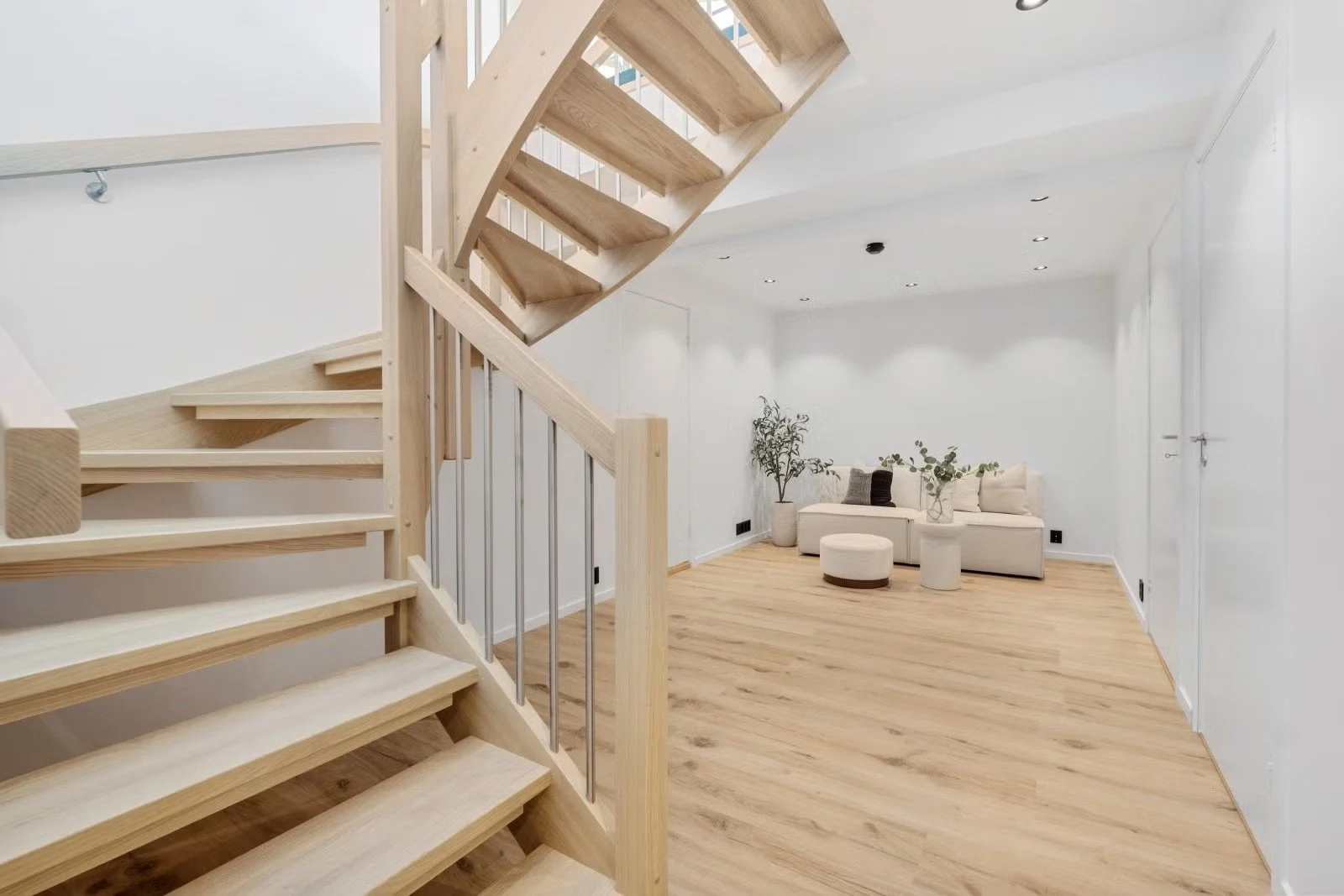 A modern interior living space with a wooden staircase on the left, light wood flooring, white walls, and a white sofa with decorative pillows in the background. There are also a few potted plants and a small round ottoman.