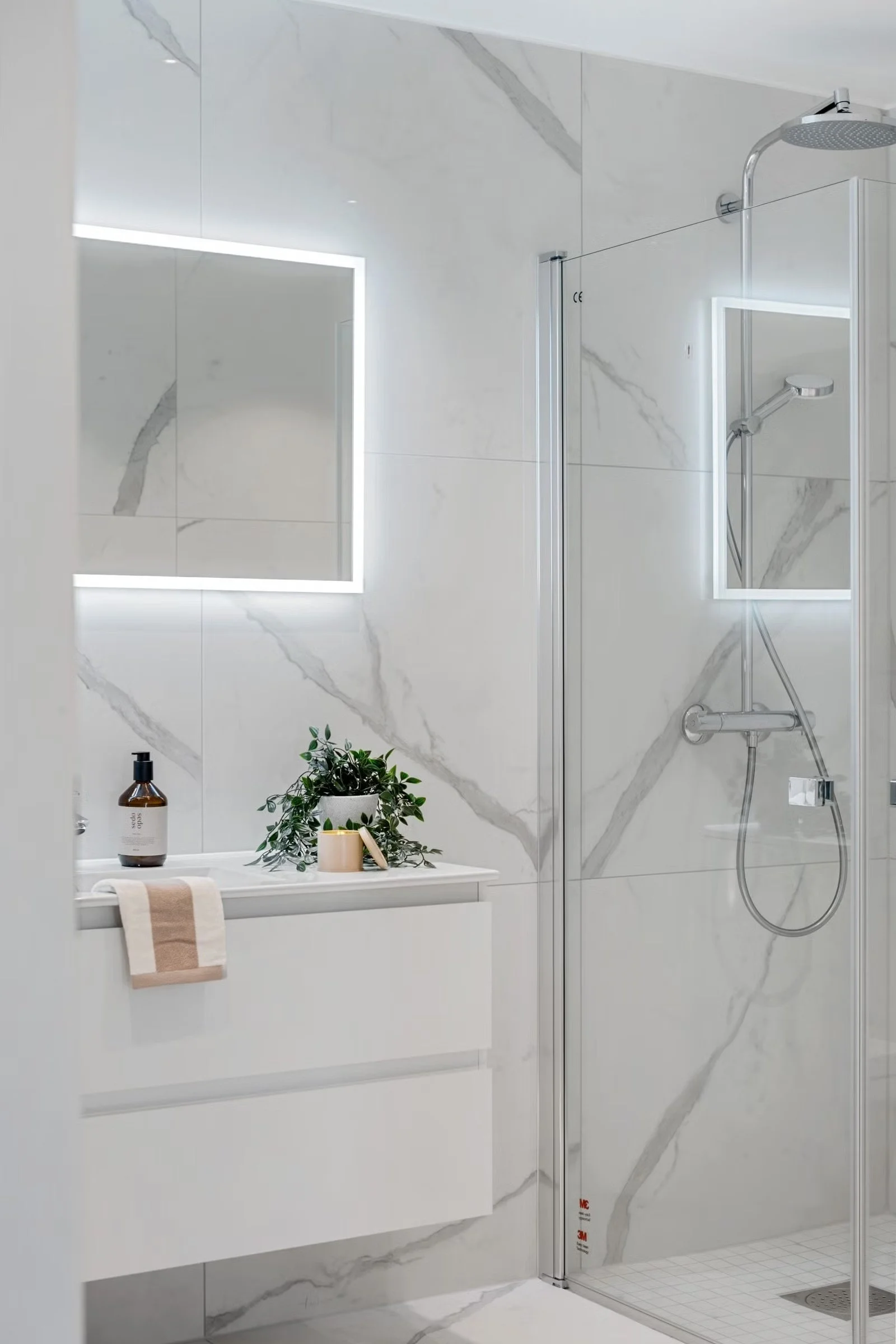 Modern bathroom with white marble tiles with gray veining, a white vanity with a beige and white towel, a mirror with built-in lighting, a small green plant, soap dispenser, and candles, and a glass-enclosed shower with a Rain showerhead.