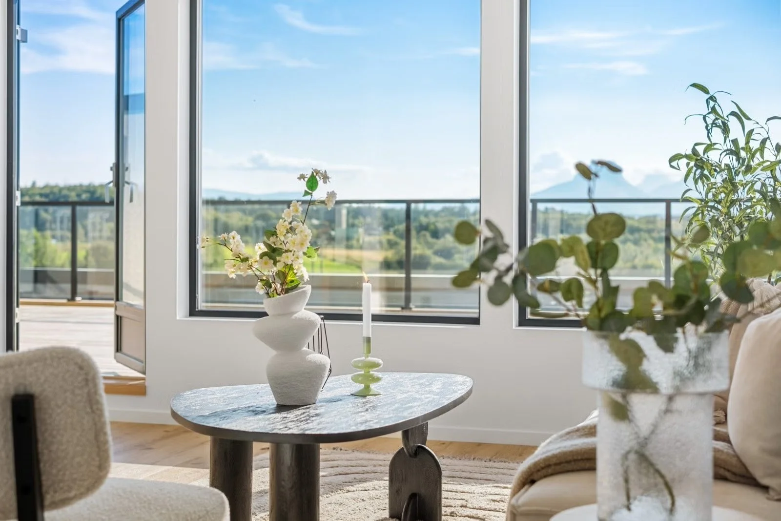 Living room with large open windows showing a view of greenery and mountains, decorated with potted plants, a table with a floral arrangement, and candles.