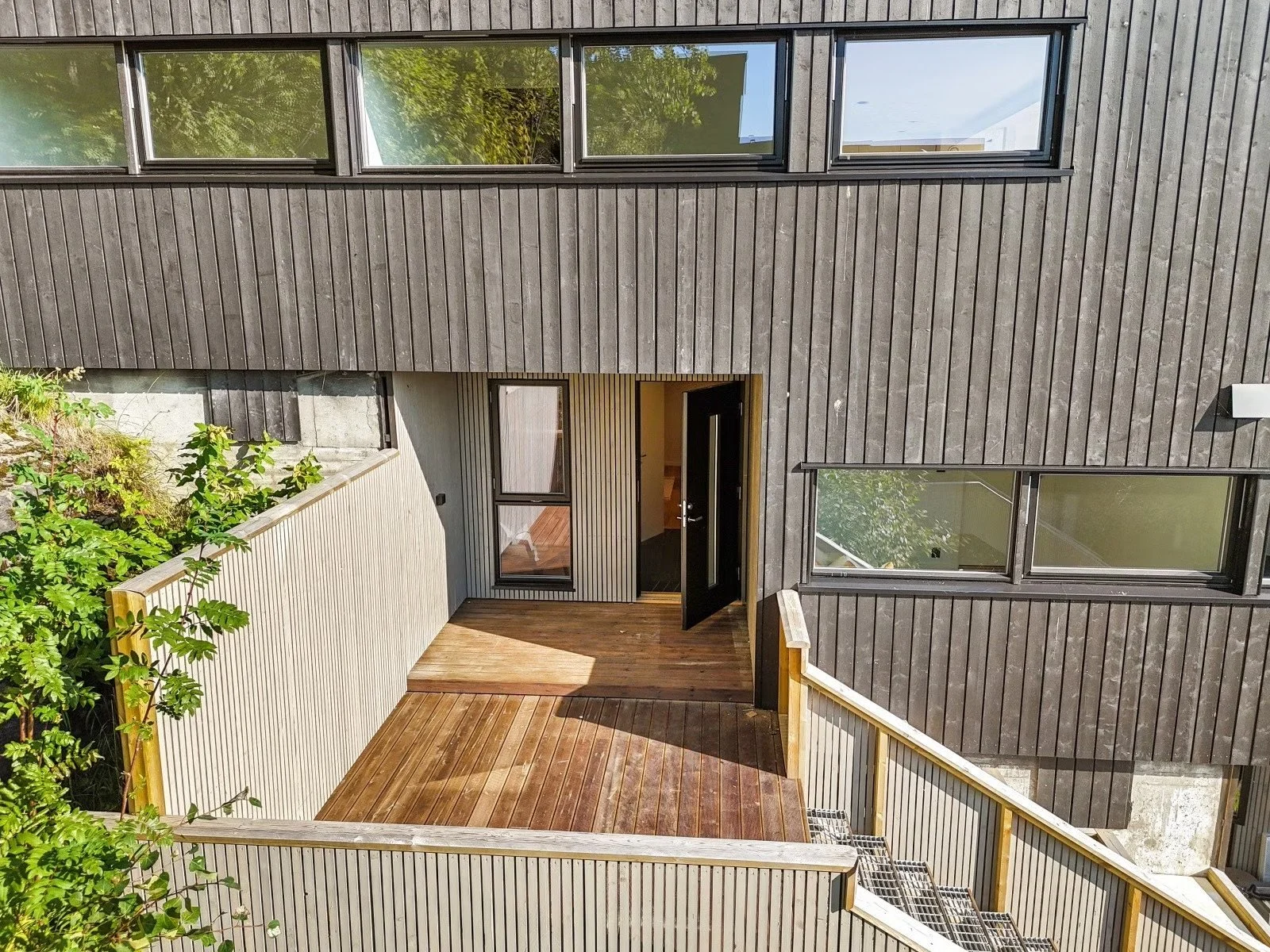 View of a modern house exterior with wooden siding, large windows, and a wooden deck entrance, surrounded by greenery.
