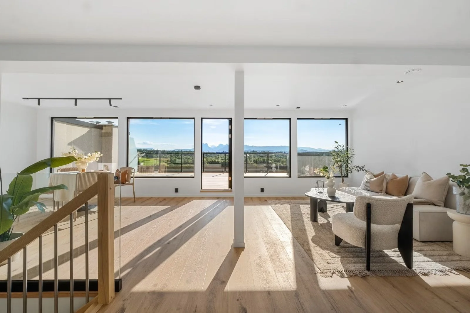 Bright living room with large windows overlooking mountains, white walls, wooden floors, and modern furniture including a beige sofa, armchair, and coffee table, decorated with plants and cushions.