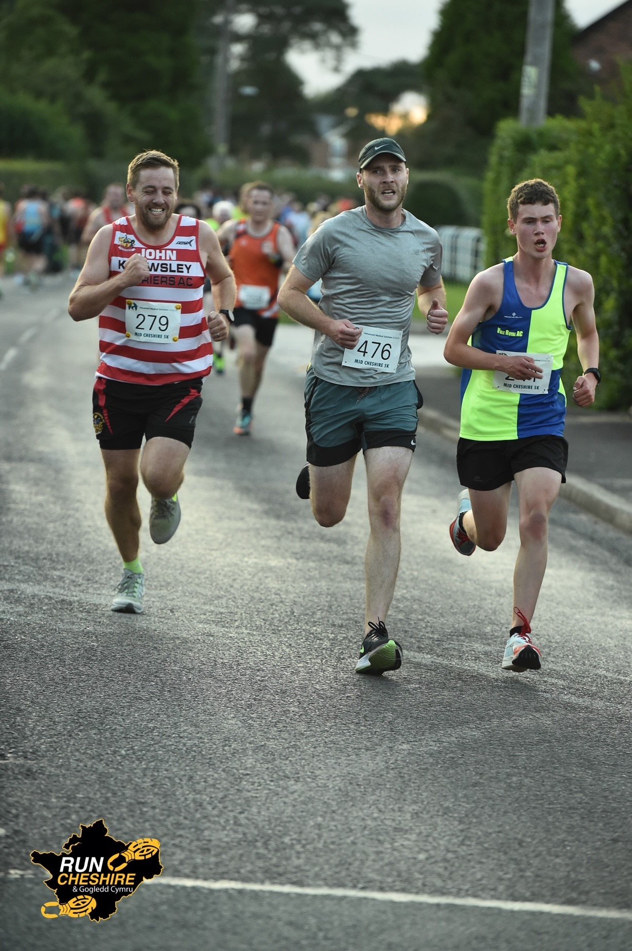 Three runners participating in the Mid Cheshire 5K race, wearing numbered bibs, running on a street with greenery and spectators in the background.