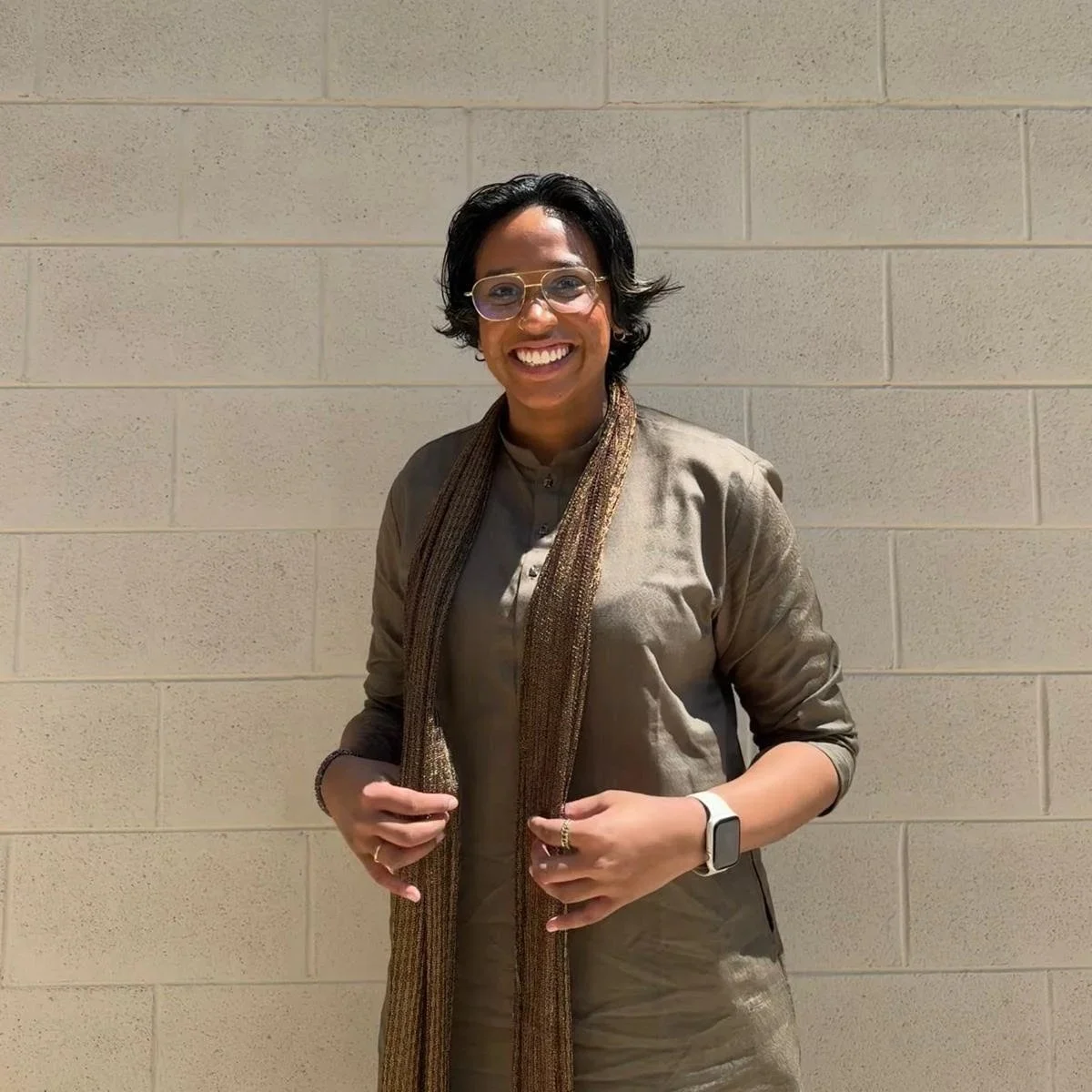 Smiling person with short dark hair, glasses, wearing a maroon suit jacket, white shirt, and patterned tie, standing indoors in front of a mirror.
