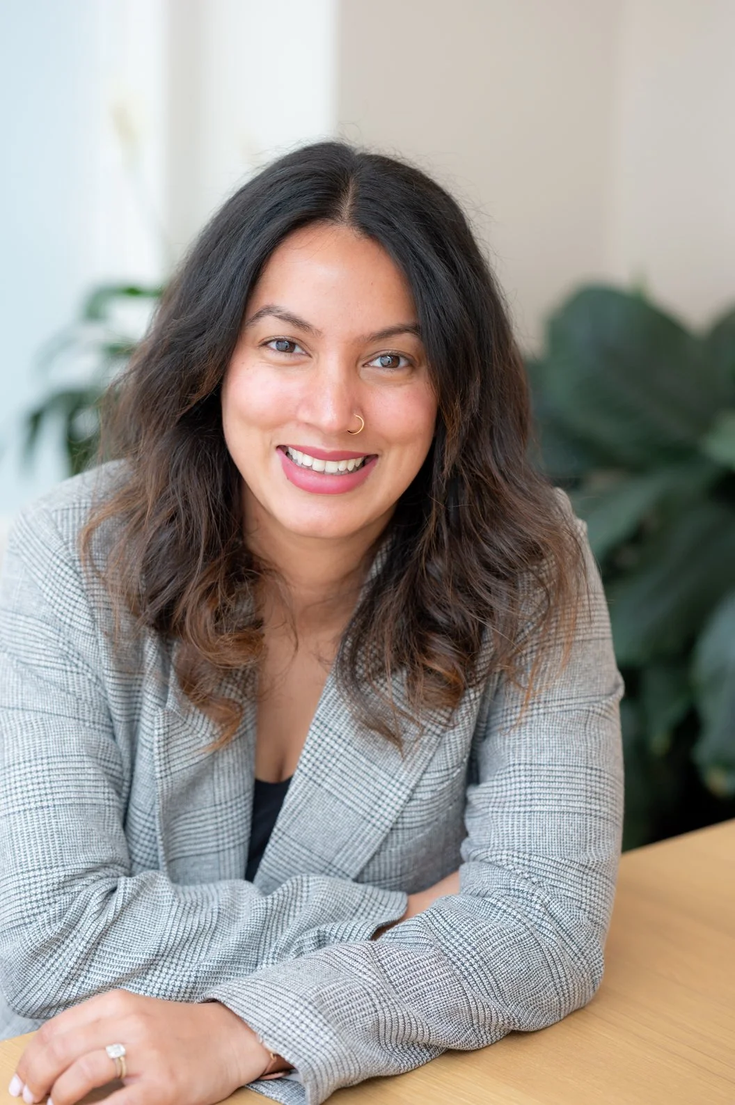 A woman in a yellow dress sits on a stone bench outdoors near the ocean, smiling at the camera with sunglasses and jewelry, with greenery, water, and a partly cloudy sky in the background.