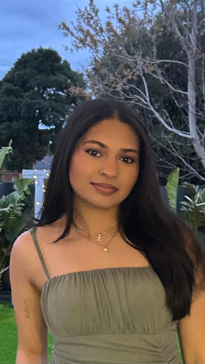 A young woman with long dark hair, wearing a khaki dress and layered necklaces, standing outdoors in a garden with trees and green plants in the background during twilight.