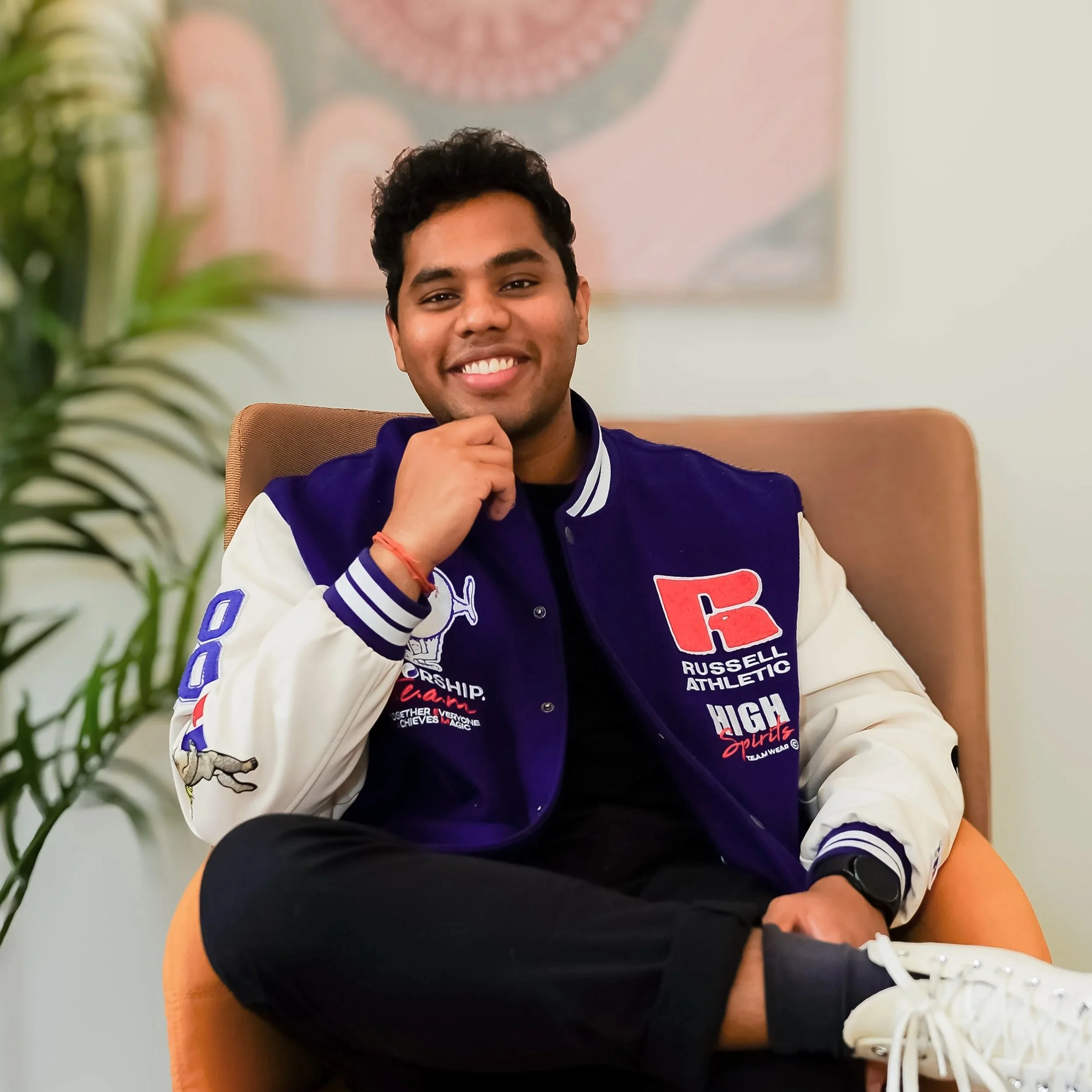 A young man with dark curly hair smiling, sitting in an orange chair, wearing a multicolored varsity jacket with various patches, black pants, and white sneakers, in a room with a plant and abstract artwork in the background.