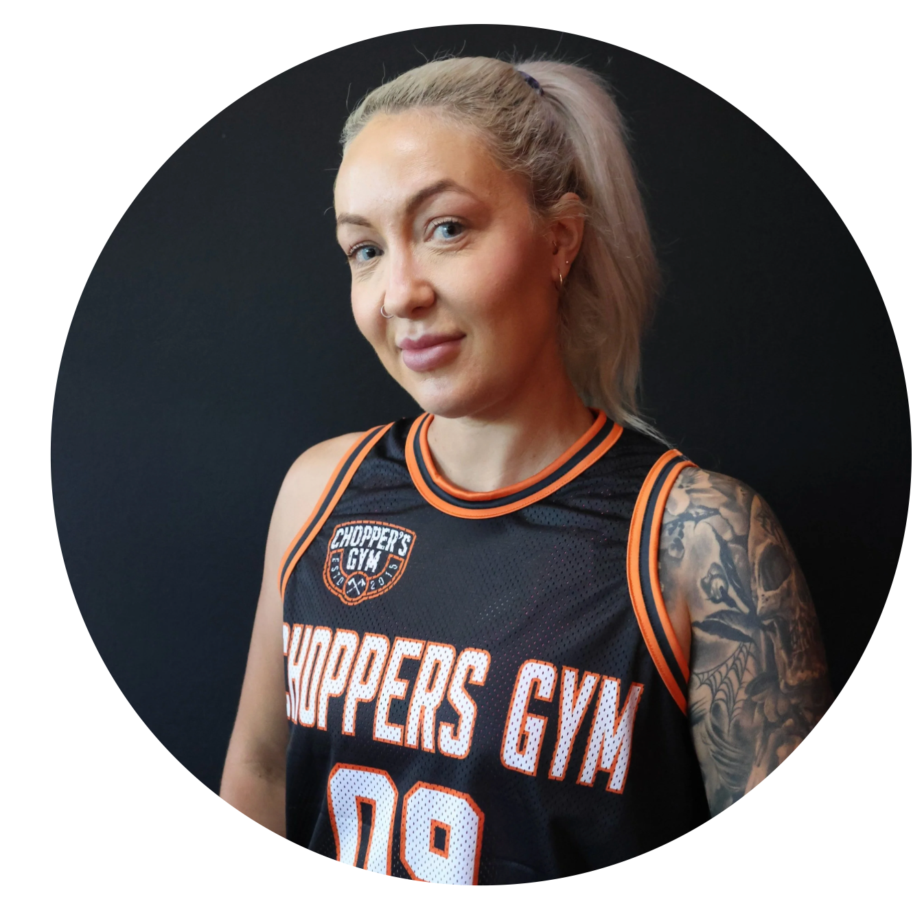 A woman wearing a black basketball jersey with orange trim that says 'Chopper's Gym' and sports a detailed tattoo on her left arm, standing against a black background.