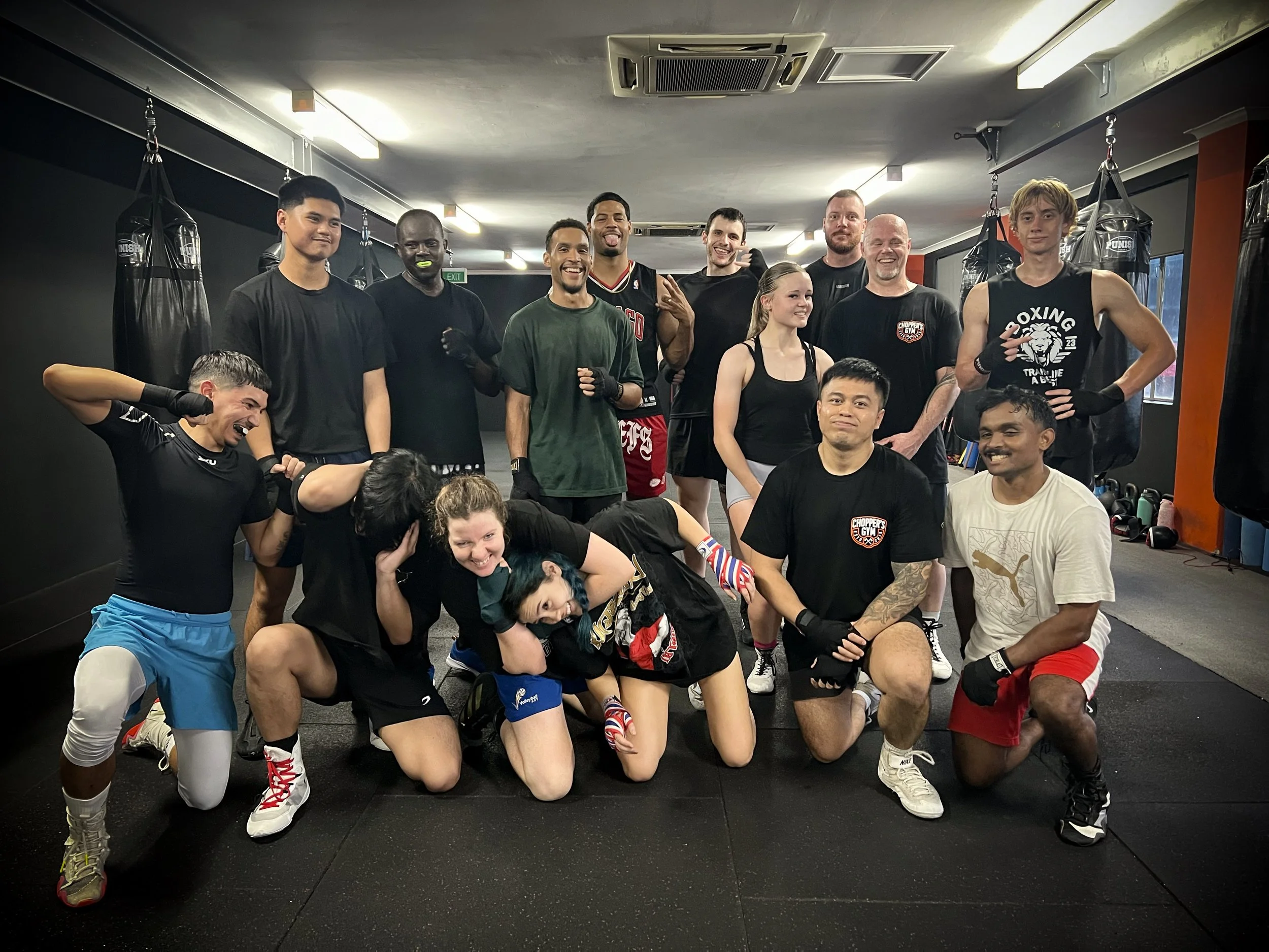 Group of people in boxing gym, posing after workout, some wearing boxing gloves and sportswear, smiling and making victory signs.