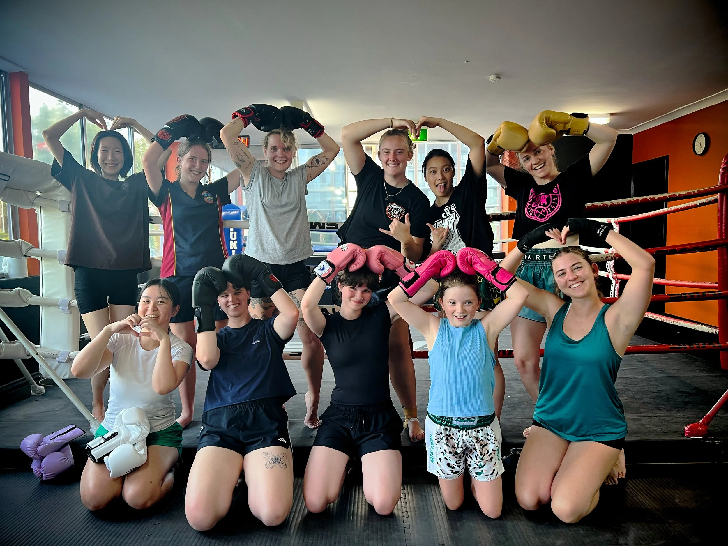 A group of diverse women and girls posing inside a boxing gym, some wearing boxing gloves and making heart shapes with their hands, smiling.