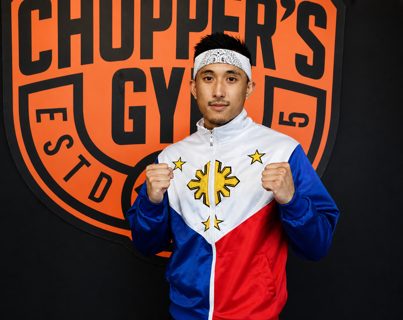 A young man in a Philippines flag-themed jacket, posing with fists raised in front of a boxing gym logo wall.
