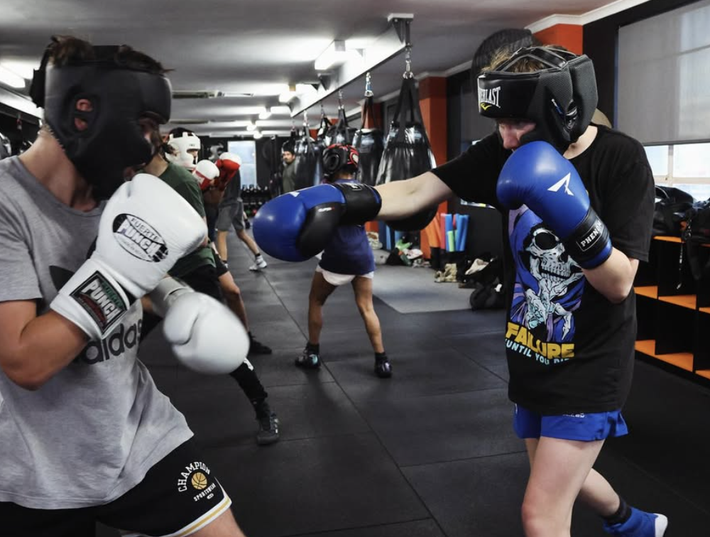 Two women practicing kickboxing in a gym, with one woman wearing a helmet and gloves, and the other delivering a punch. Several people are seen in the background.