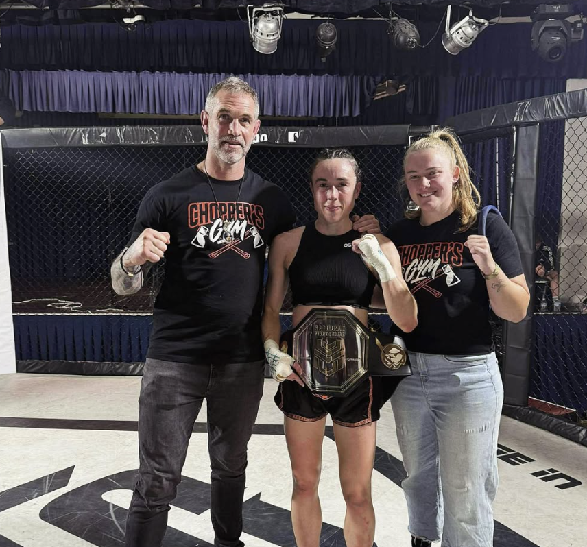 Group of individuals kneeling in a row during a martial arts class, with their instructor standing in front of them. The participants are wearing boxing gloves and athletic clothing in a gym with punching bags hanging from the ceiling.