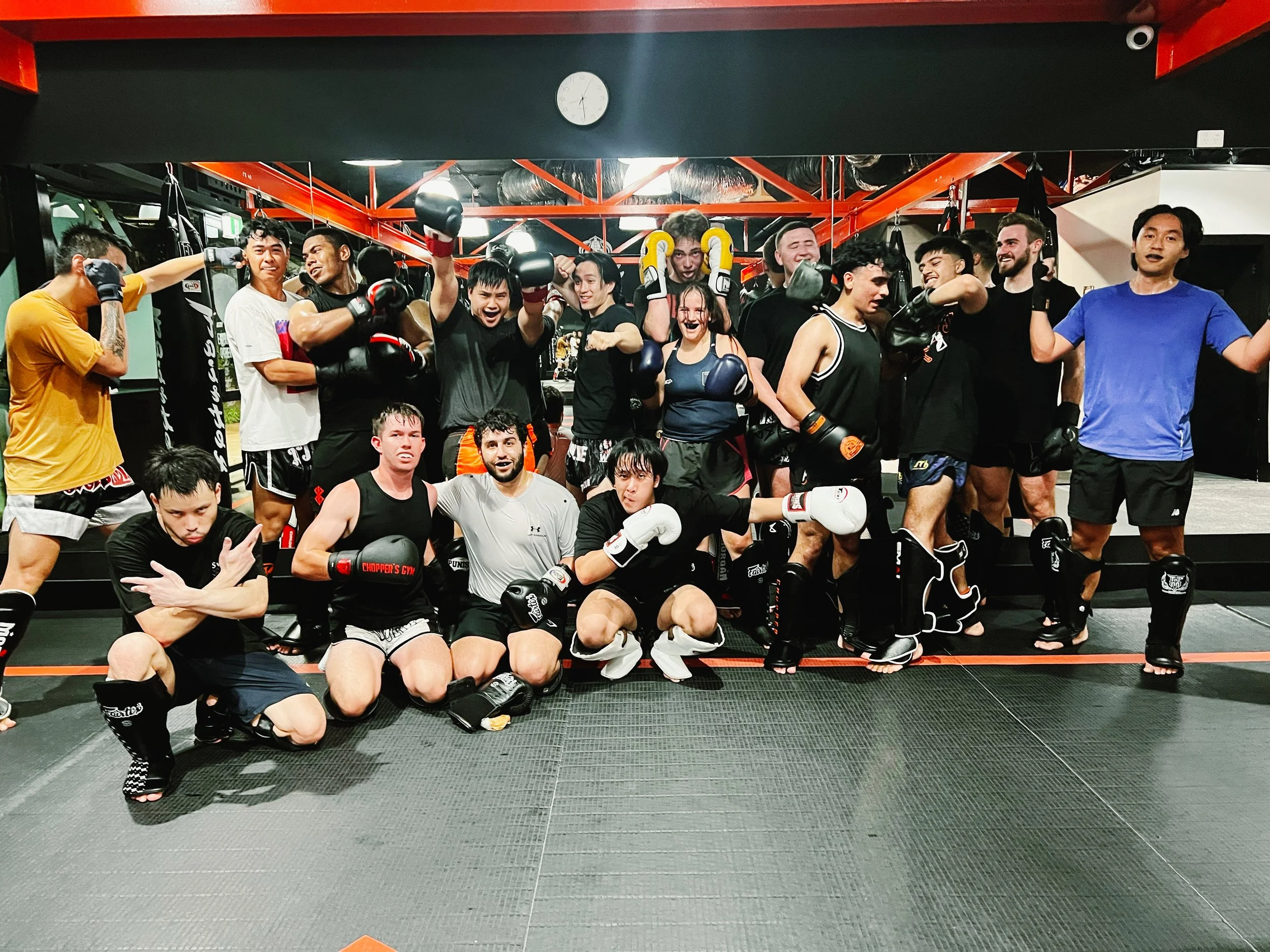 Group of diverse people wearing mixed martial arts gear, pose, and celebrate in a gym with boxing bags and a clock showing 7:15.