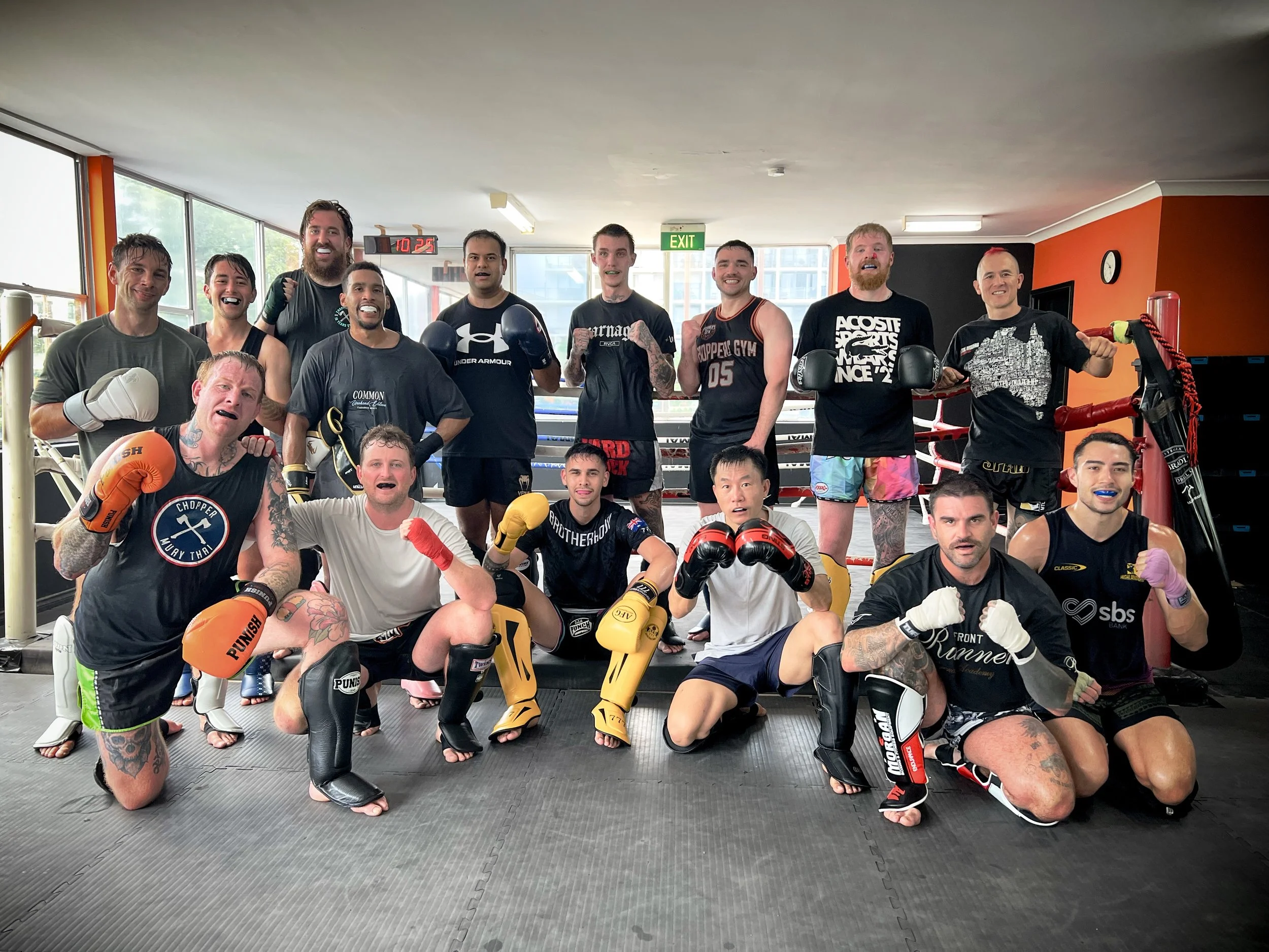 Group of diverse martial artists and fighters posing together in a gym training area. Some wearing boxing gloves and MMA gear, showing readiness for training or competition.