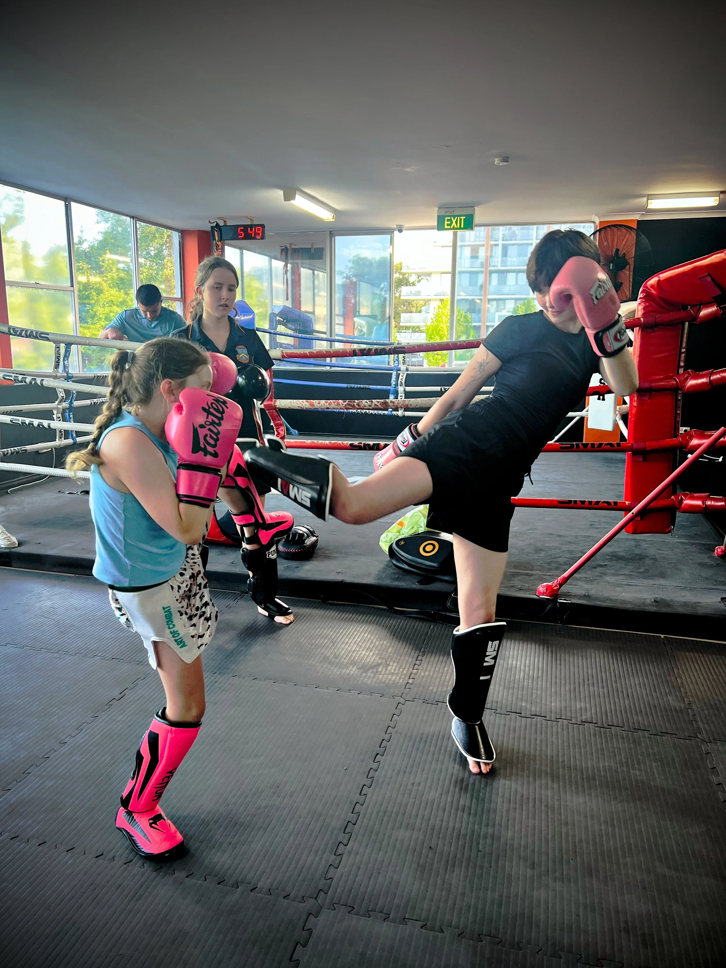 Two young women practicing kickboxing in a gym with boxing gloves, one in pink gear and the other in black gear, while a girl watches. There is a boxing ring, large windows, and a digital clock showing 5:49.