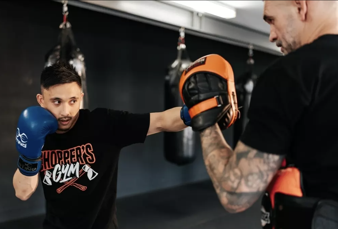 Young man practicing boxing with an older trainer in a gym, wearing boxing gloves and throwing a punch at a punching pad held by the trainer.