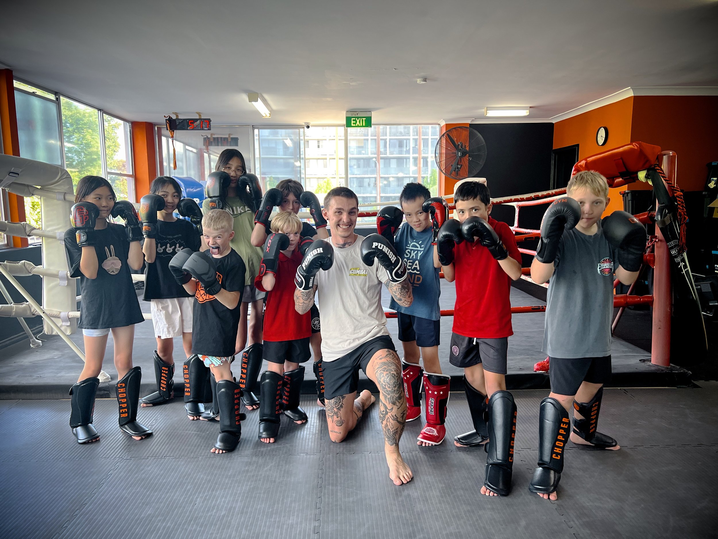 Group of children and an adult martial arts instructor inside a gym, all wearing boxing gloves and martial arts gear, posing in fighting stances.