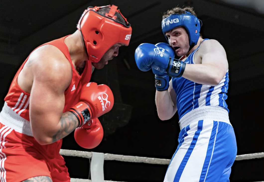 Group of individuals kneeling in a row during a martial arts class, with their instructor standing in front of them. The participants are wearing boxing gloves and athletic clothing in a gym with punching bags hanging from the ceiling.