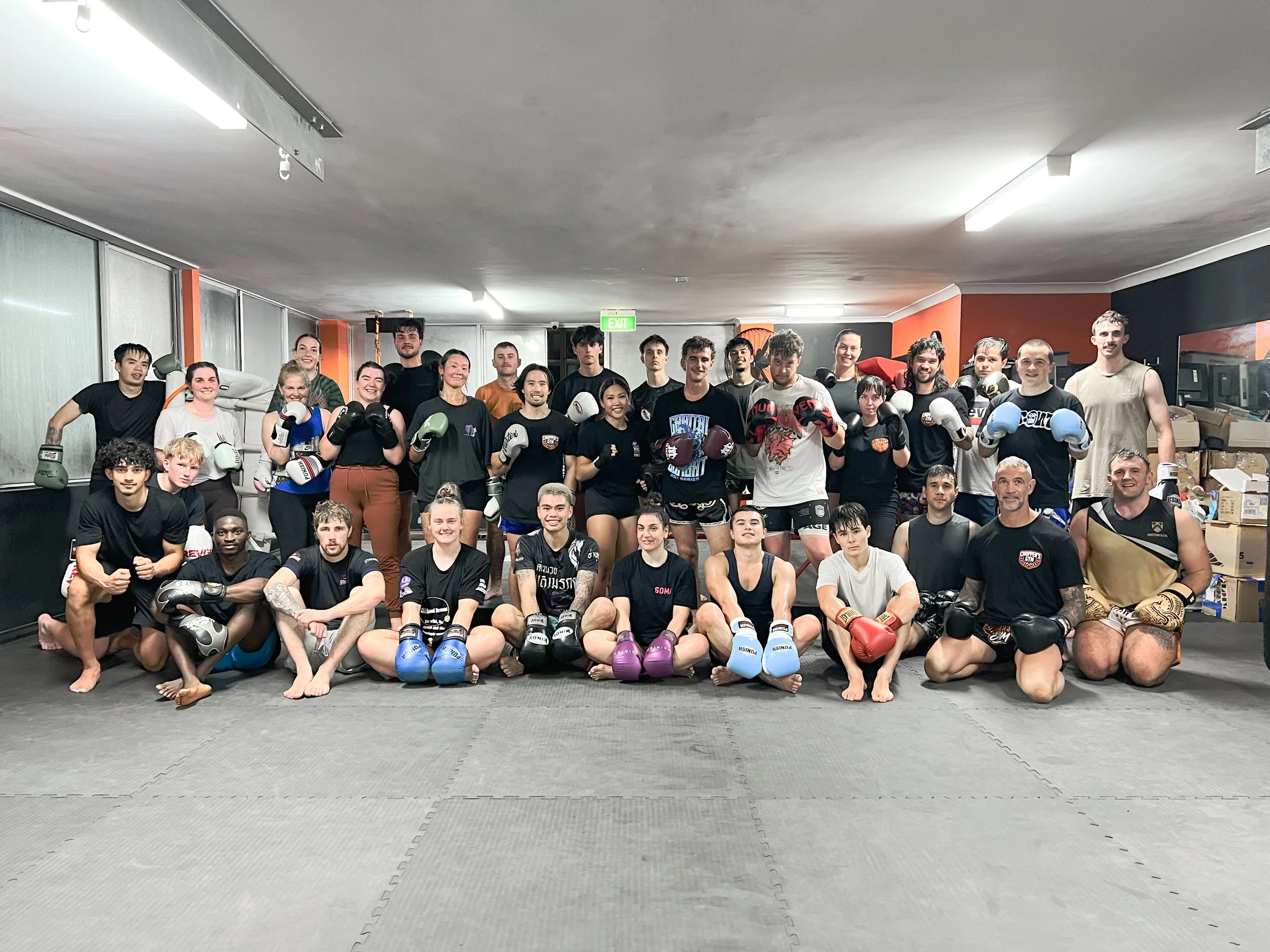 Group of martial arts or boxing students, consisting of children, teenagers, and adults, posing for a photo in a training facility, some wearing boxing gloves.
