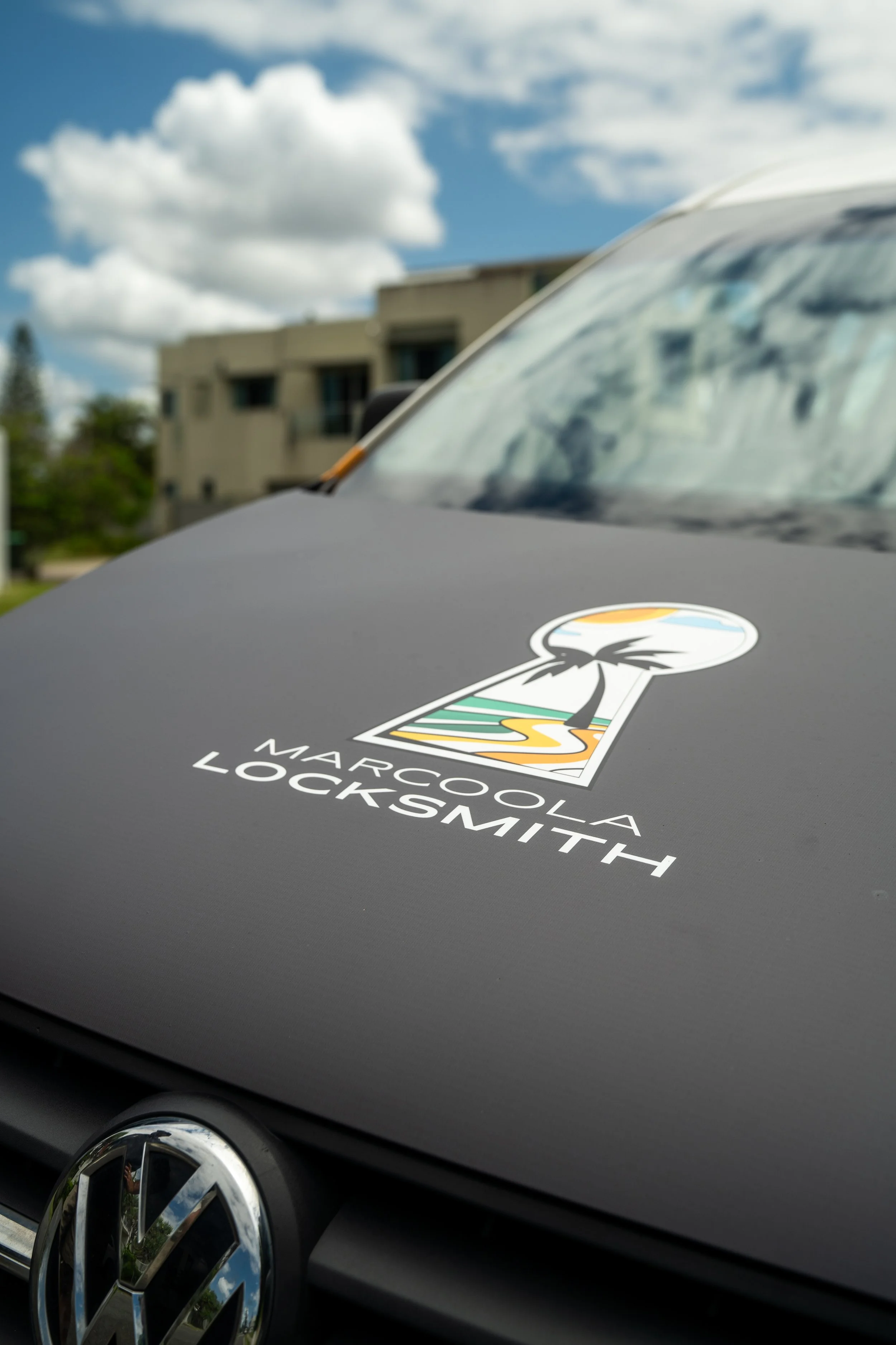 Close-up of the front of a black Volkswagen car with a "Marocoola Locksmit" sticker on the hood, and the Volkswagen logo visible on the grille. The background shows a modern residential building and a cloudy sky.