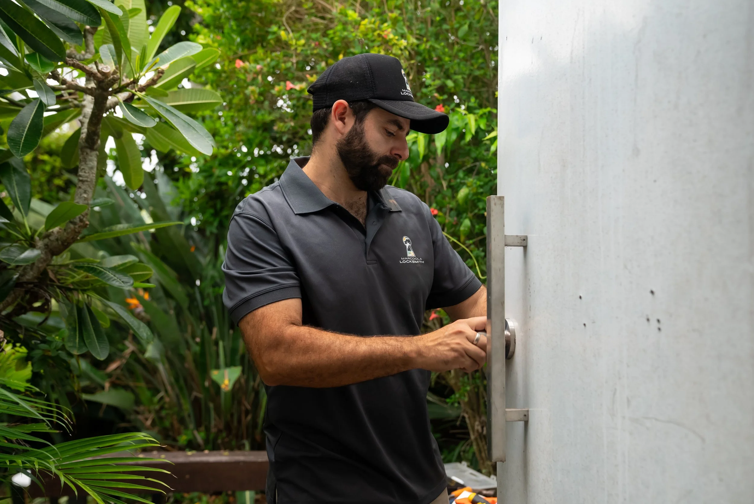 A man wearing a black cap and a gray polo shirt labeled 'Marcola Locksmith' is unlocking a white door with a silver handle, standing outdoors among green shrubbery.