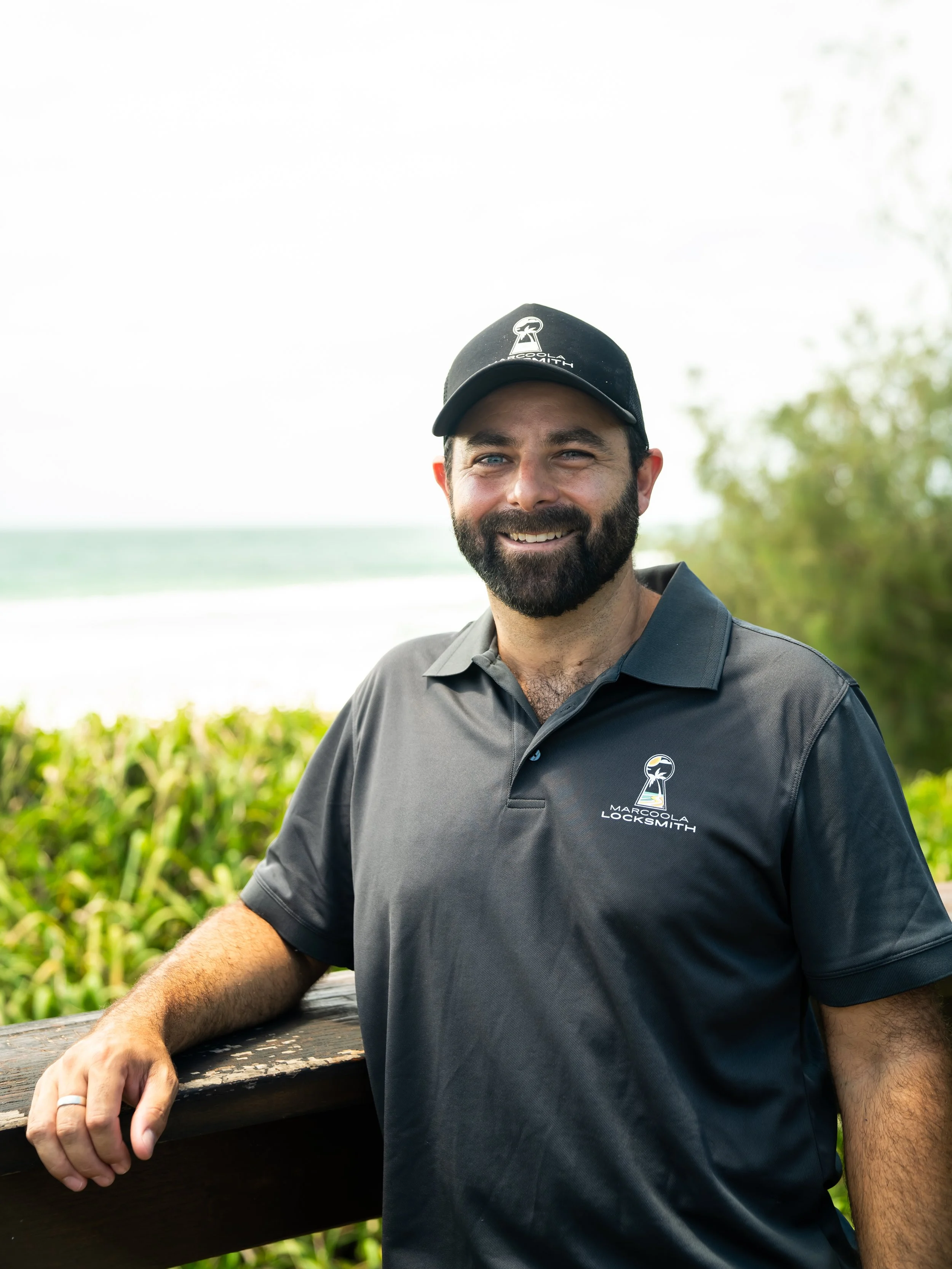 Smiling man with a beard wearing a black cap and a black polo shirt with a logo that says 'Marcoola Locksmith', sitting outdoors on a wooden railing with greenery and a cloudy sky in the background.