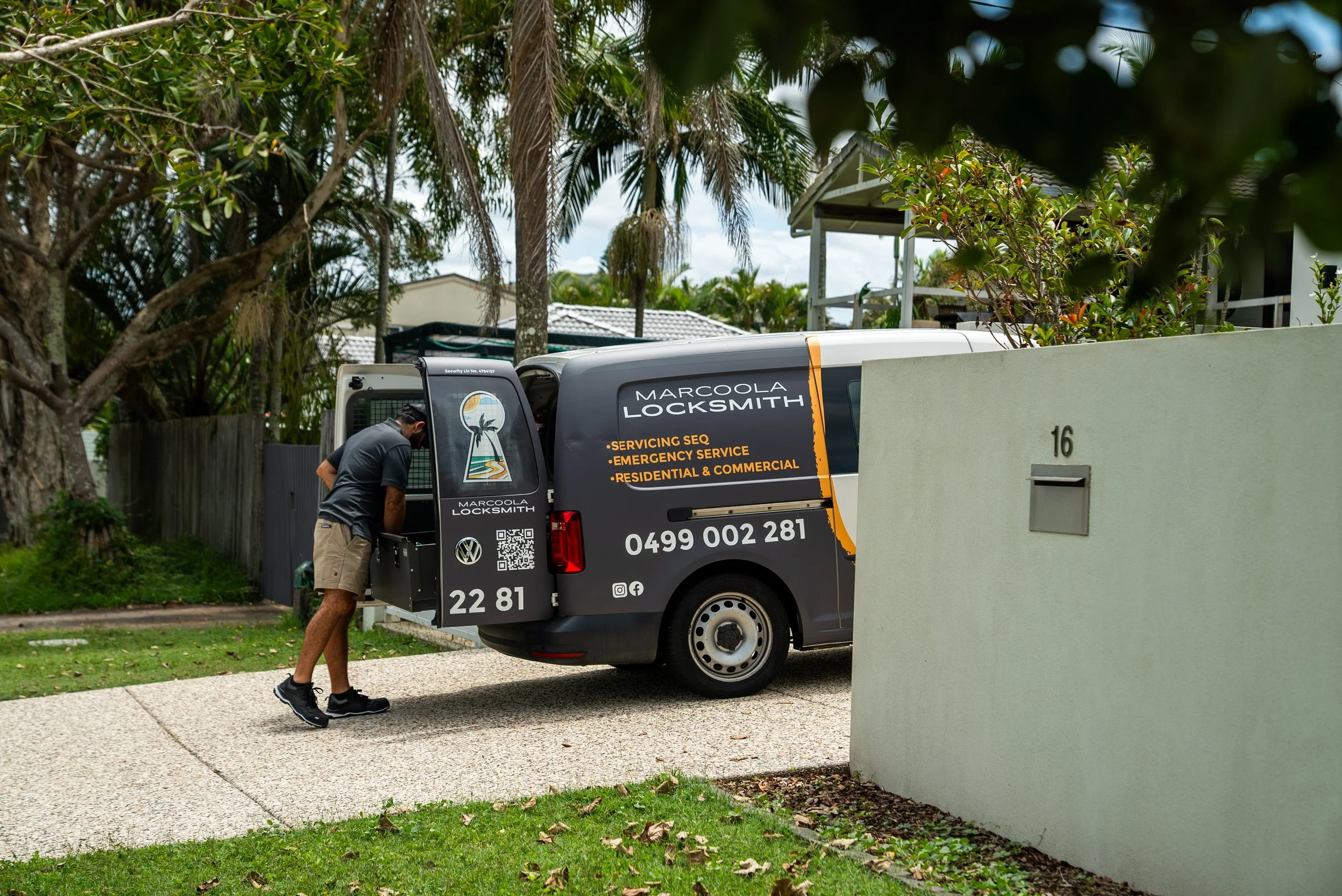 A man is working on a locksmith van parked in a suburban area with trees and houses nearby. The van has company information and services listed on it.