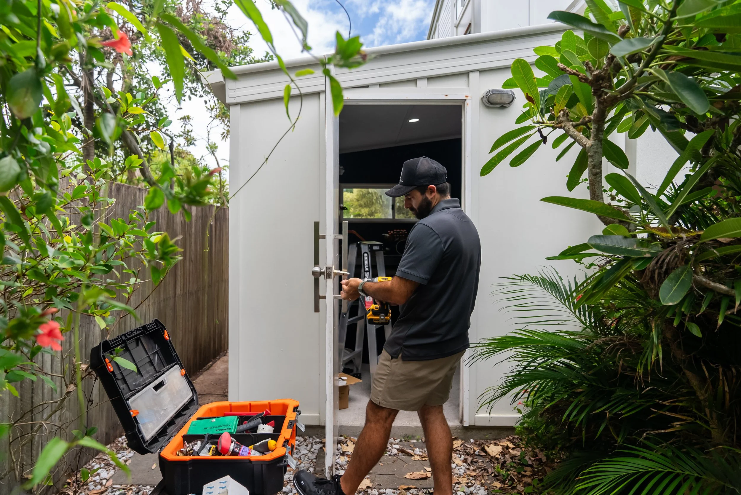 A man working on an outdoor shed surrounded by lush green plants, with an open toolbox and tools on the ground.