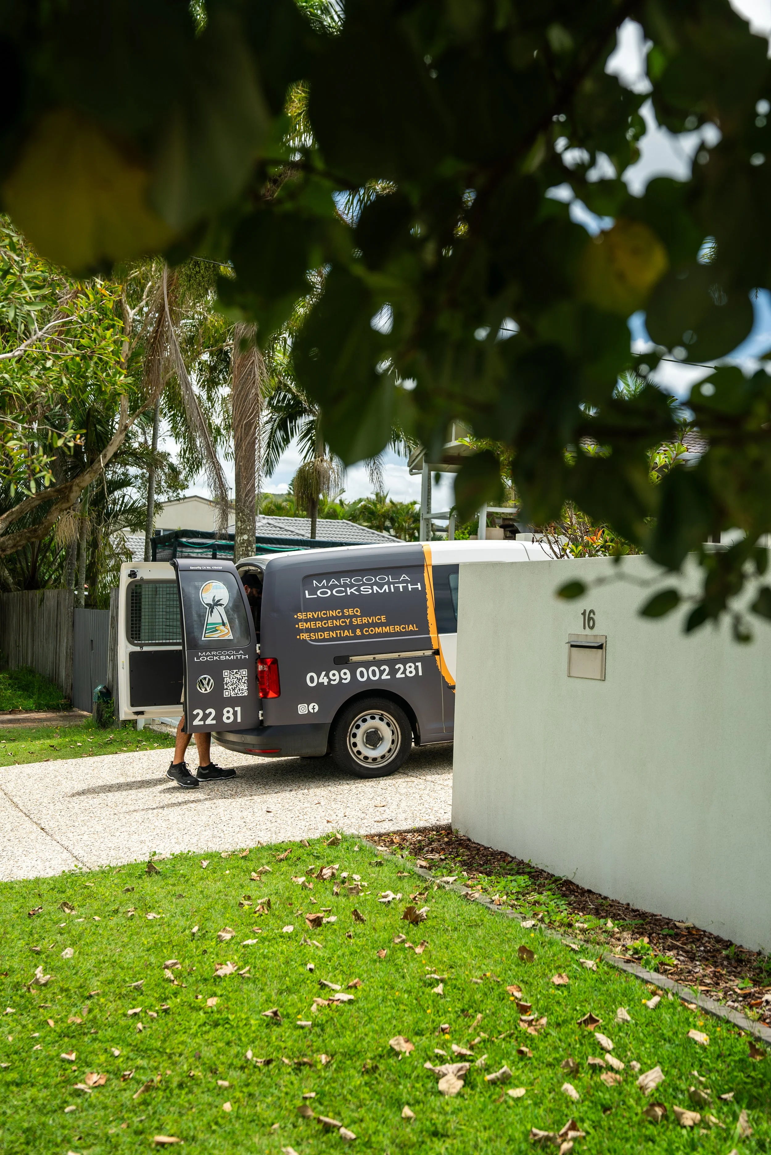 A locksmith worker is about to enter a van labeled 'Marcola Locksmith' which offers services like servicing security, emergency, residential, and commercial. The van is parked in a residential area with a green lawn and trees, and a tall white wall partially obstructs the view.