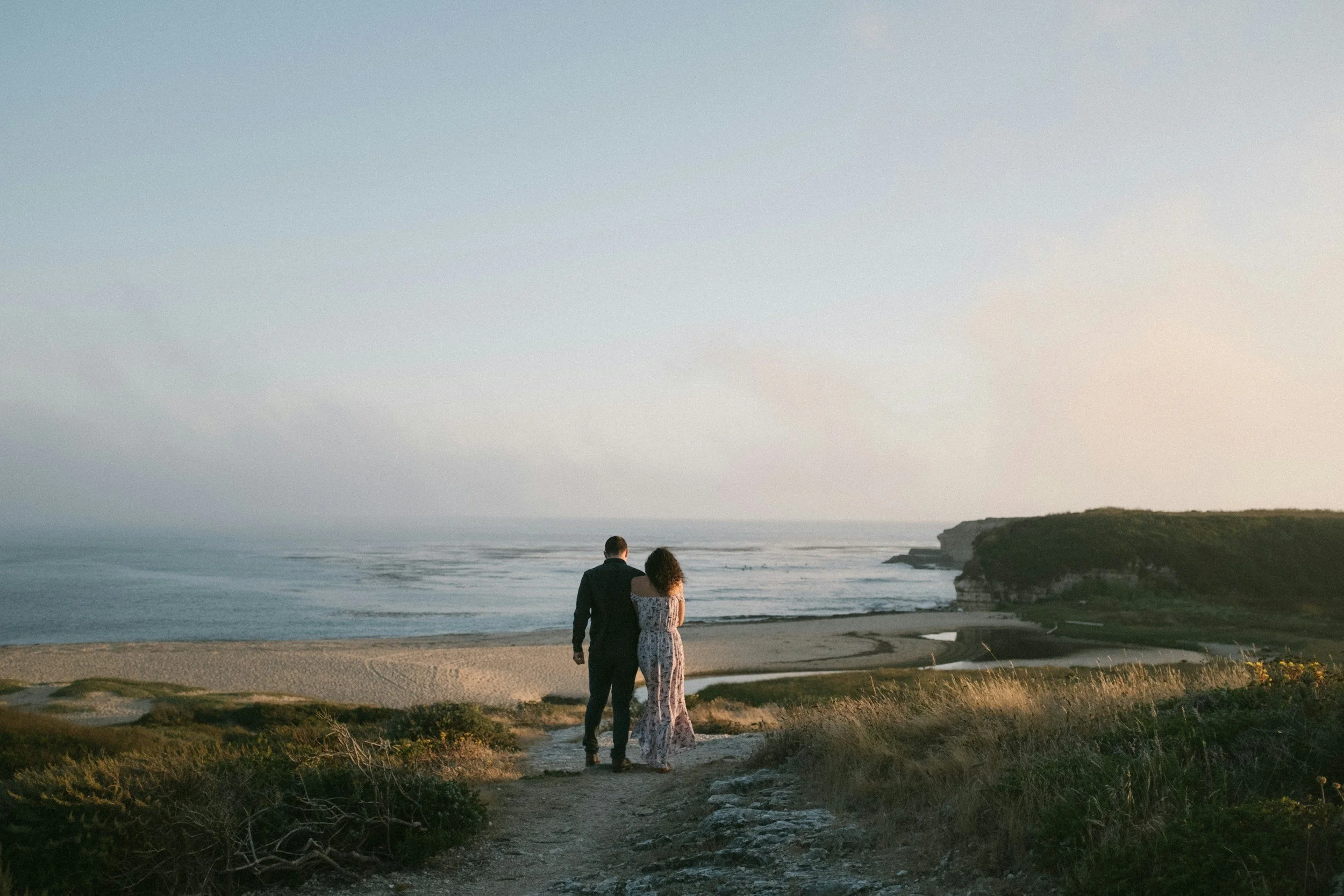 A couple walking towards a scenic coastline, surrounded by grassy fields and overlooking the ocean. It's a serene and picturesque landscape at sunset.