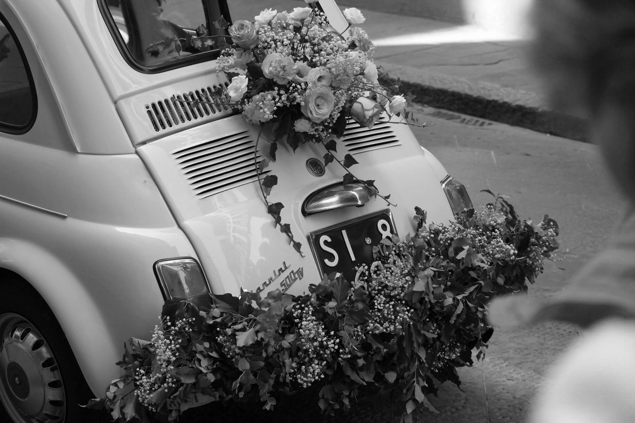 Vintage car decorated with flowers, parked on a street. The car's rear is adorned with floral arrangements, including roses and greenery.