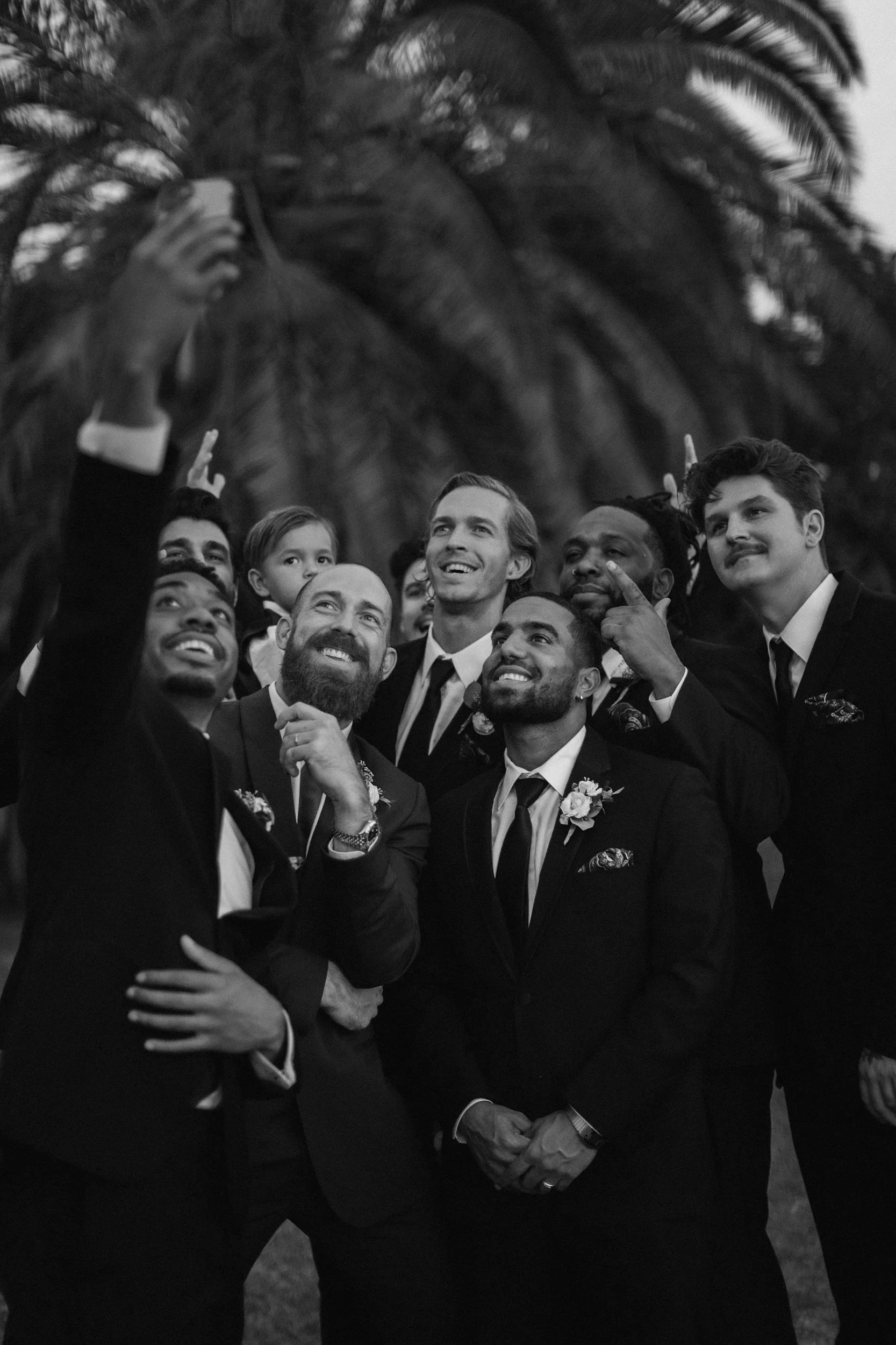 Group of men in suits taking a selfie outdoors with palm trees in the background.