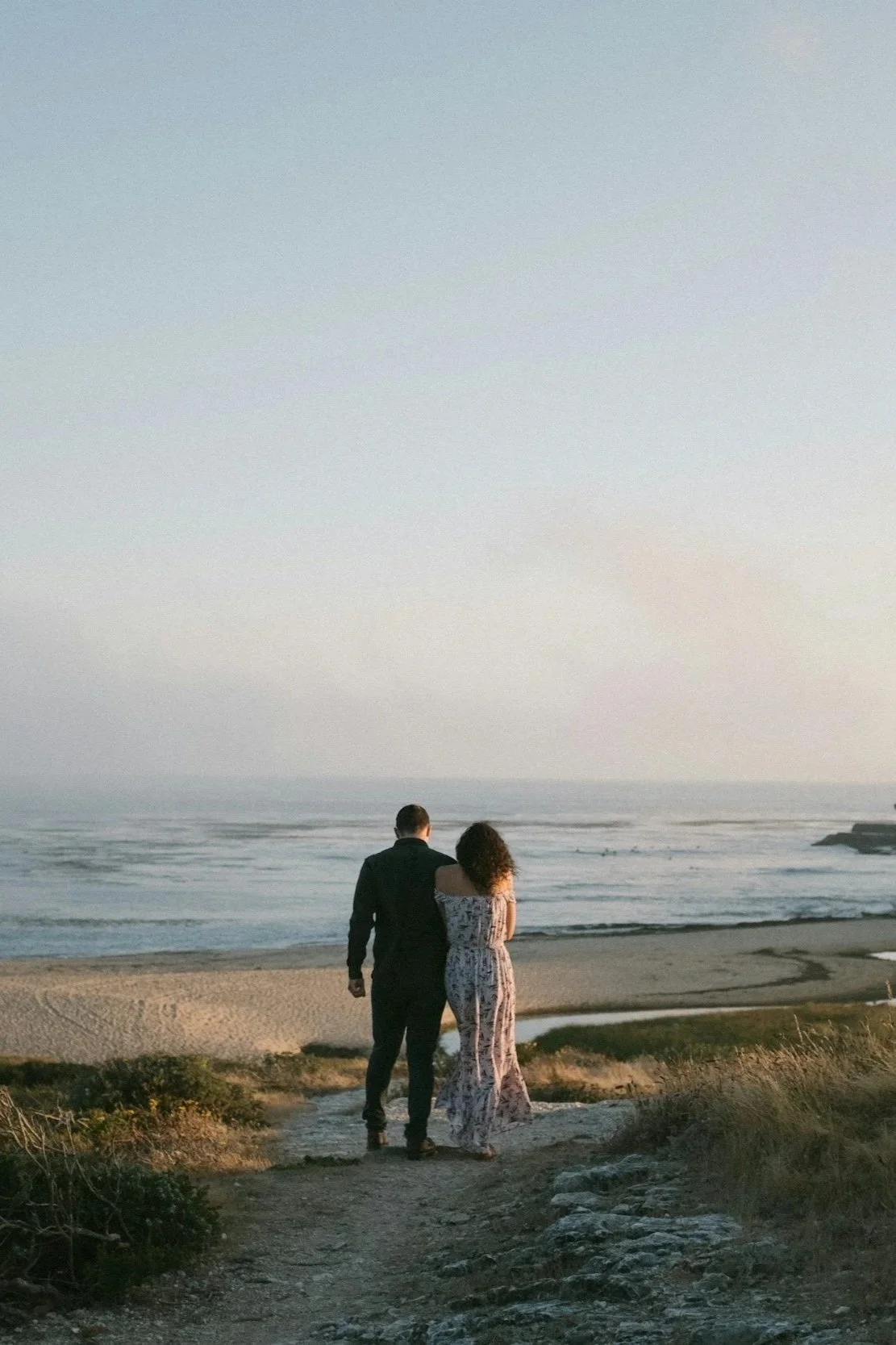 A couple walking towards a scenic coastline, surrounded by grassy fields and overlooking the ocean. It's a serene and picturesque landscape at sunset.