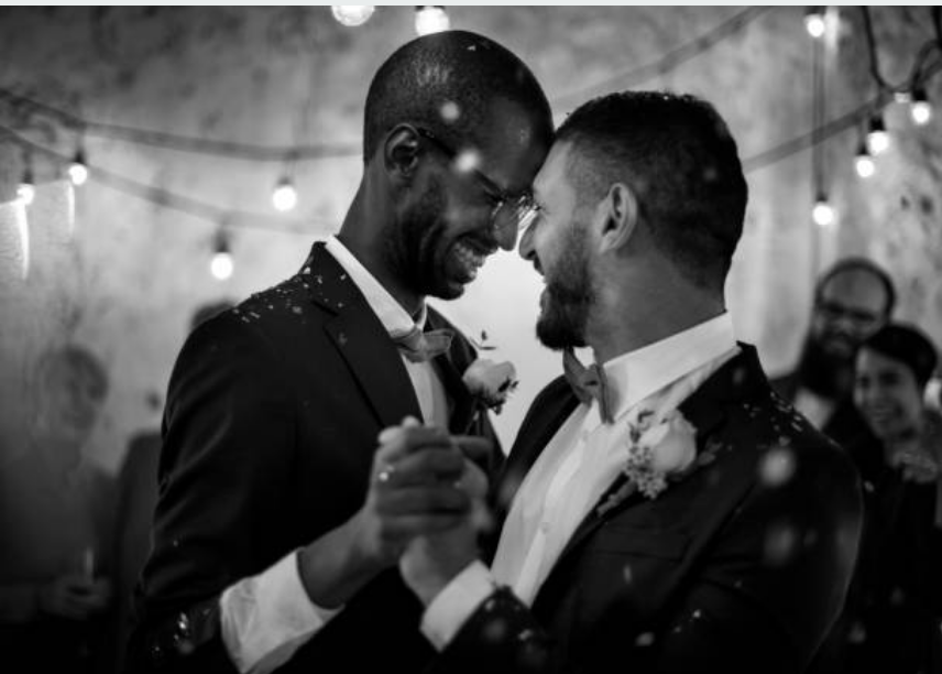 Homosexual Wedding, LGBTQI+, LGBTQ Wedding, Two men in suits dancing at a wedding, smiling at each other, with people in the background and string lights overhead.