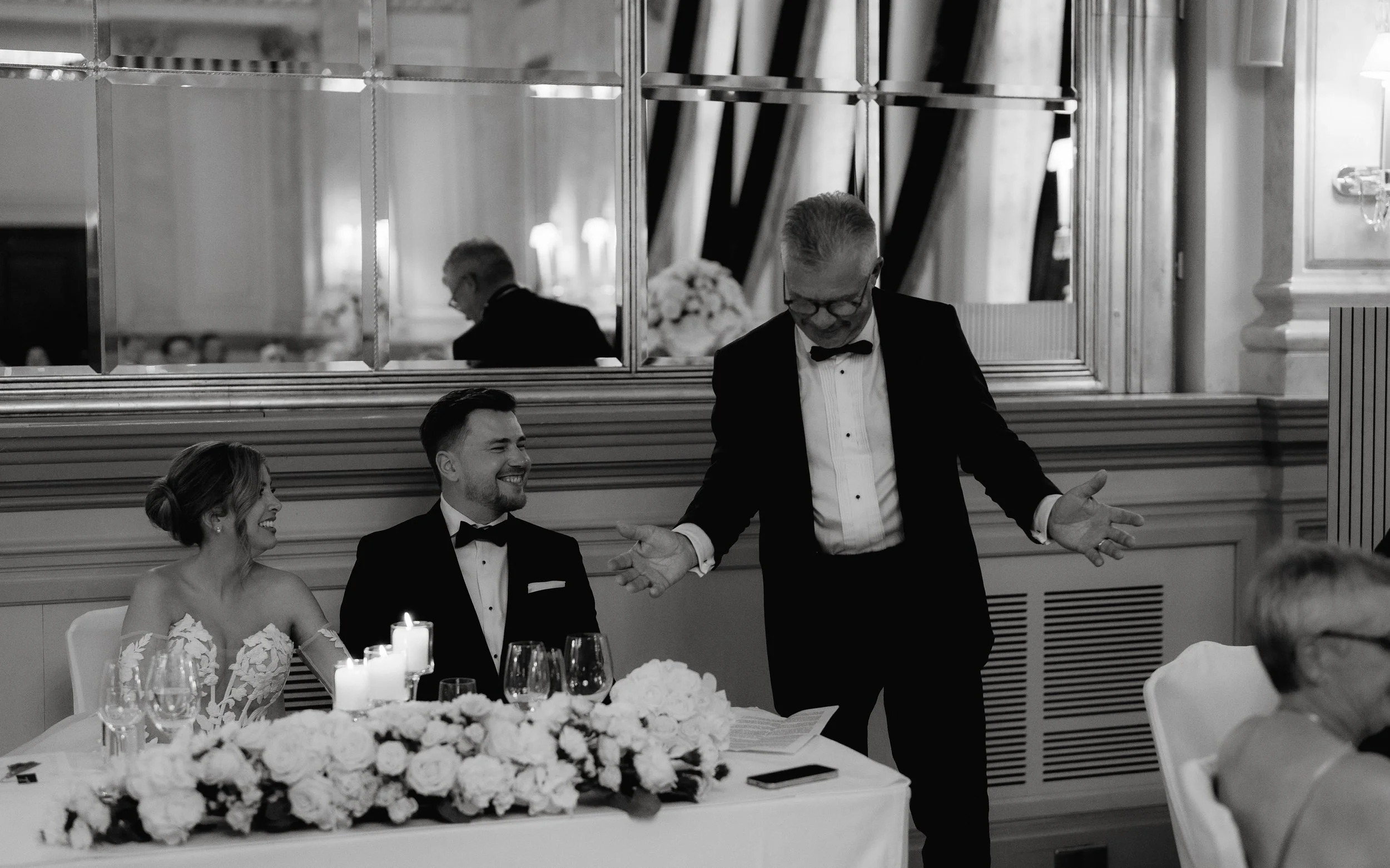 Black and white photo of a wedding speech scene with a bridal couple and a suited man.