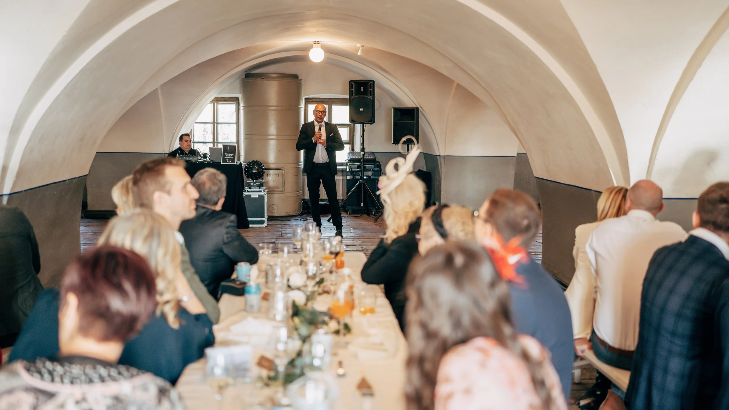 Man speaking at an indoor event, guests seated at tables under arched ceiling