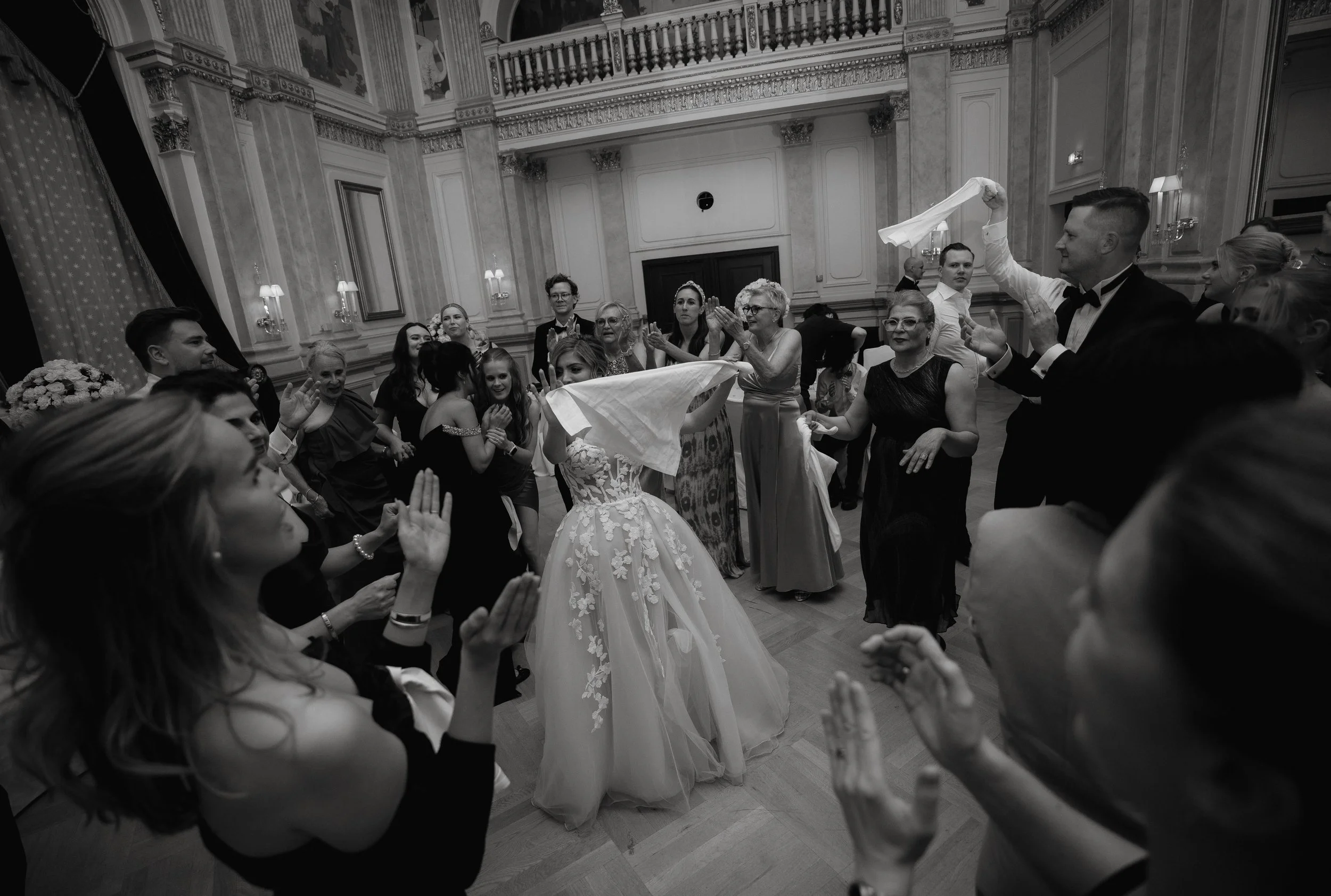 Black and white photo of a wedding celebration with people dancing and clapping around a bride in a formal gown.