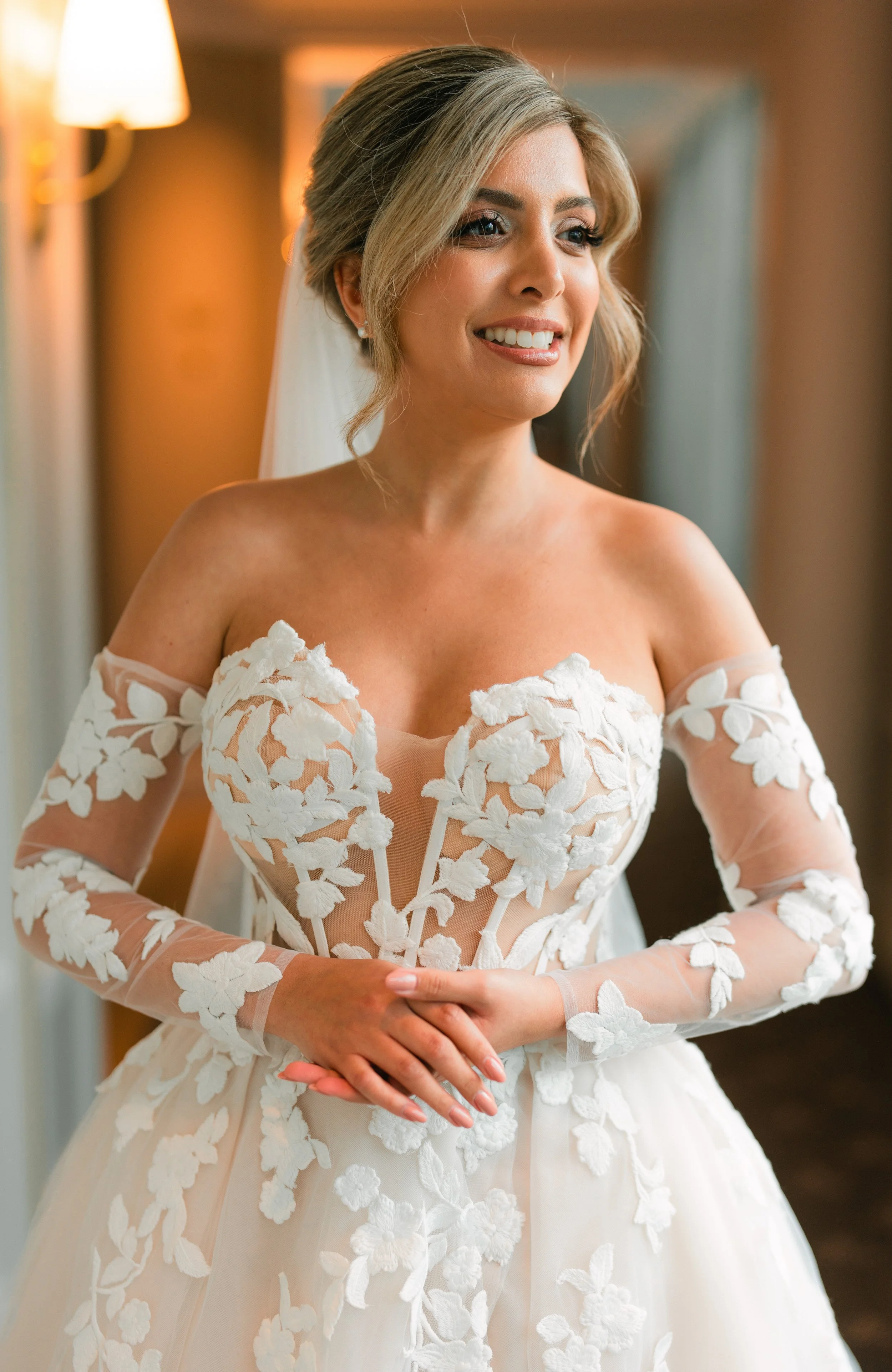 Bride in white floral wedding dress, smiling and standing indoors.