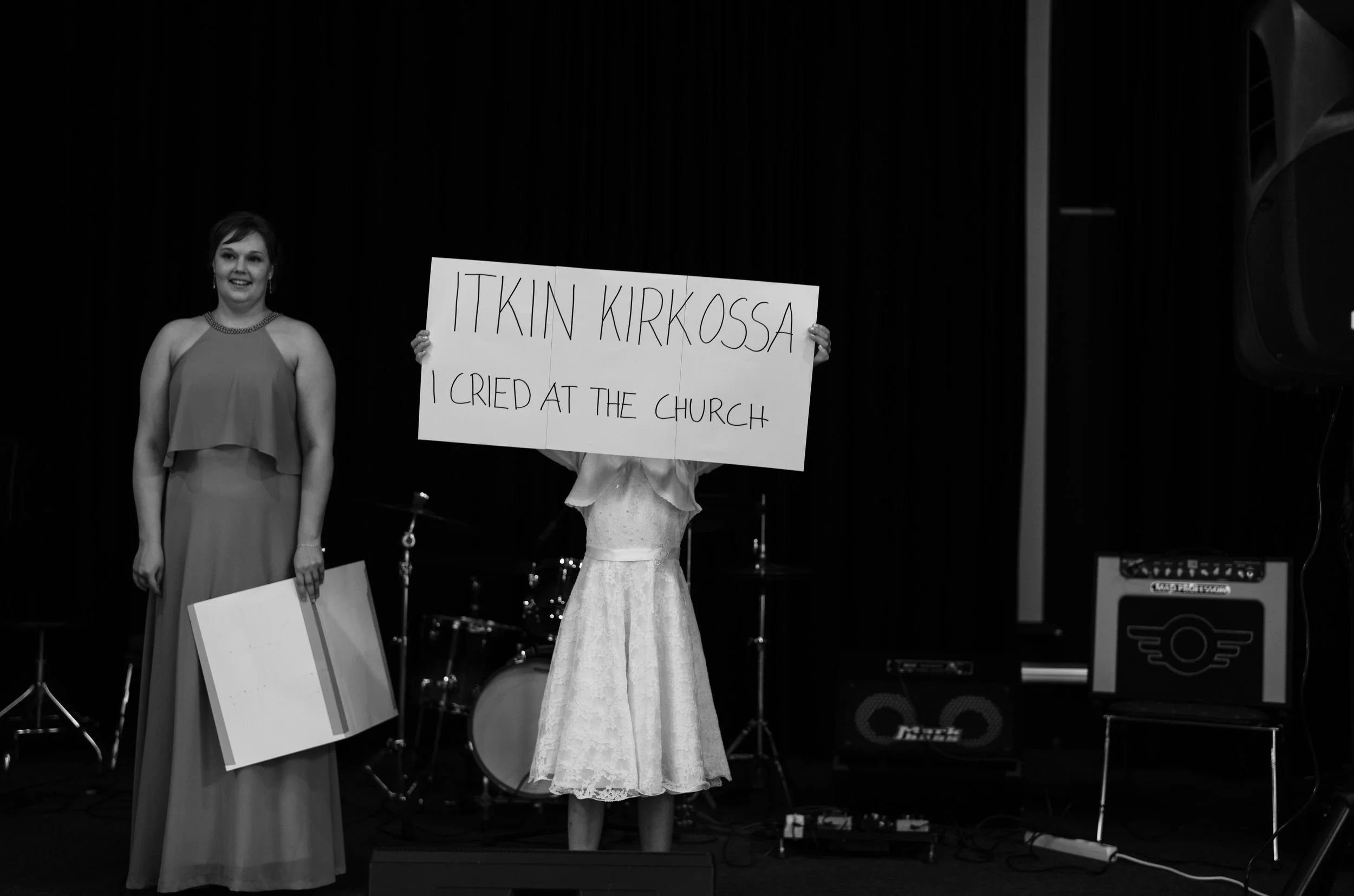 Black and white photo of two people on stage; one holding a sign reading 'ITKIN KIRKOSSA I CRIED AT THE CHURCH'.