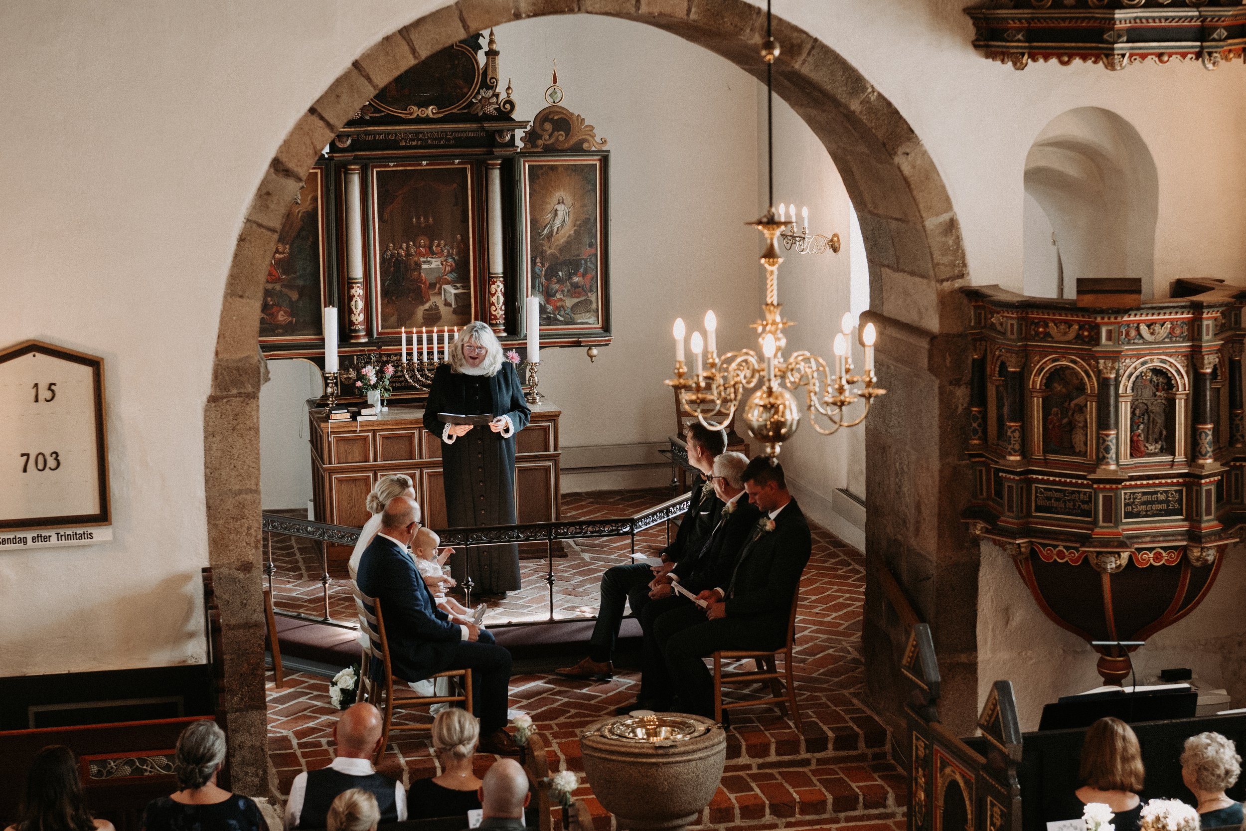 Interior of a church with a pastor officiating a wedding ceremony. Guests are seated, and decorative elements like a chandelier and ornate pulpit are visible.