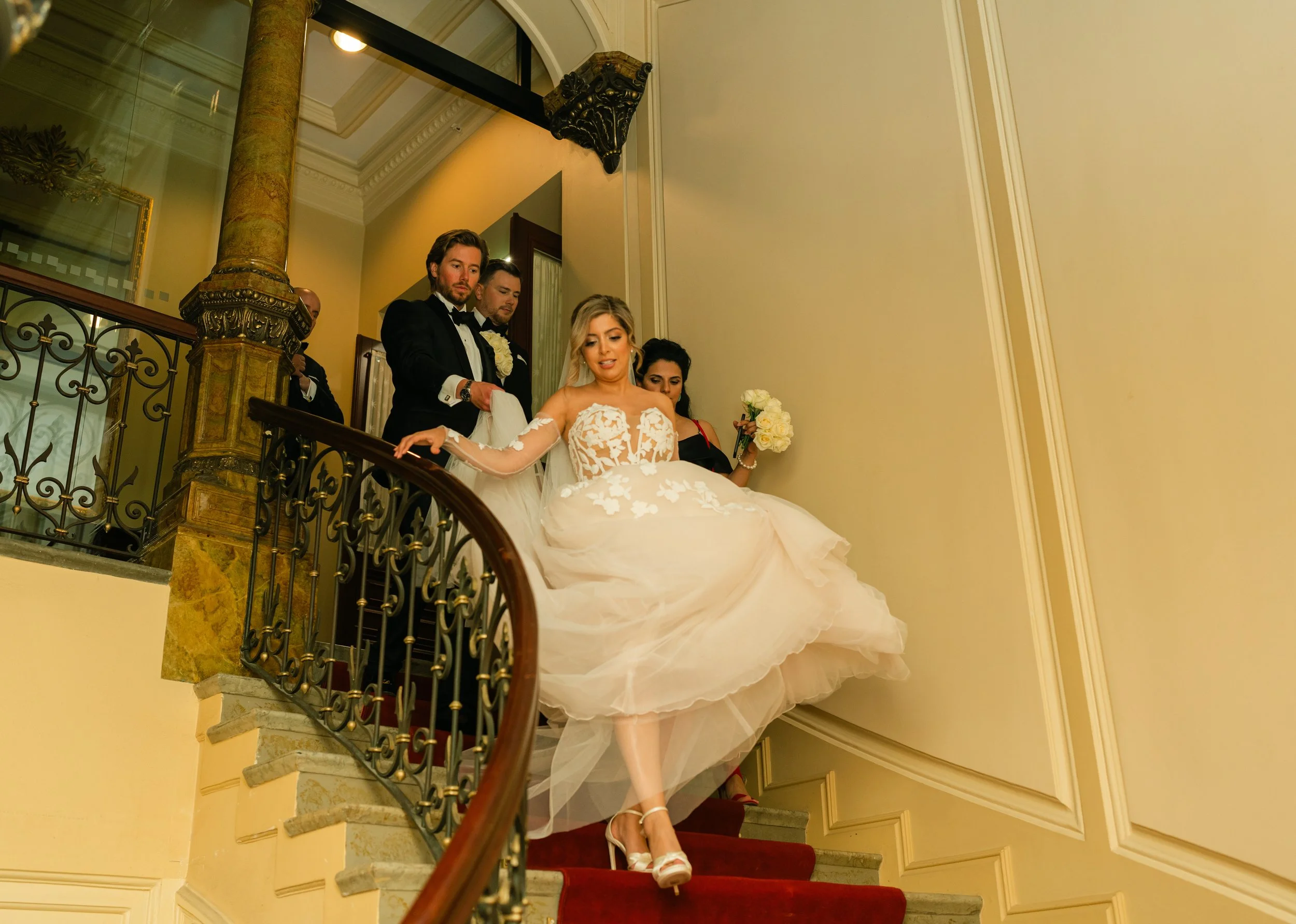 Bride descending staircase with bridal party, elegant setting, wedding attire.