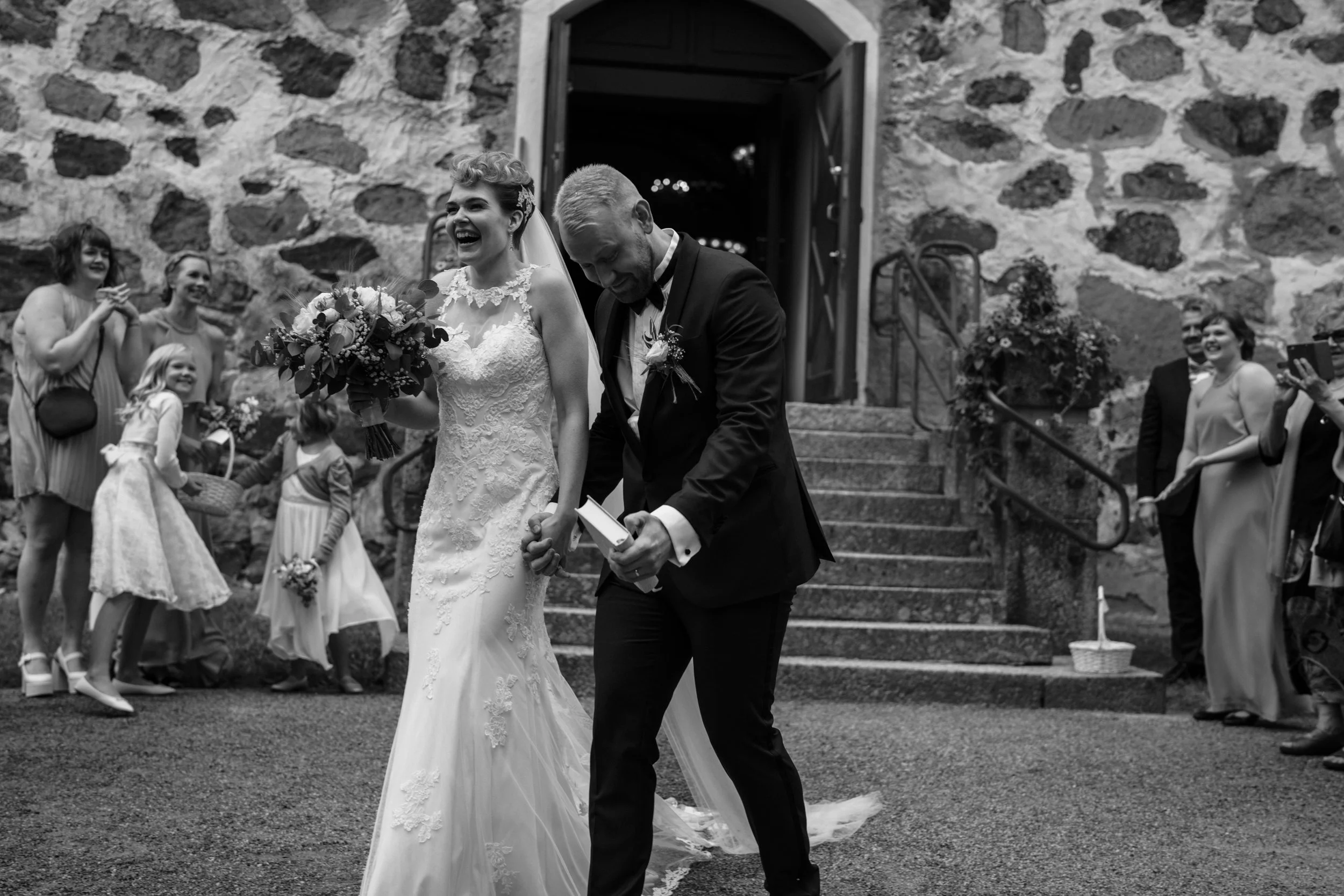 Bride and groom laughing outside church after wedding ceremony, holding hands, surrounded by guests and children, black and white photo.