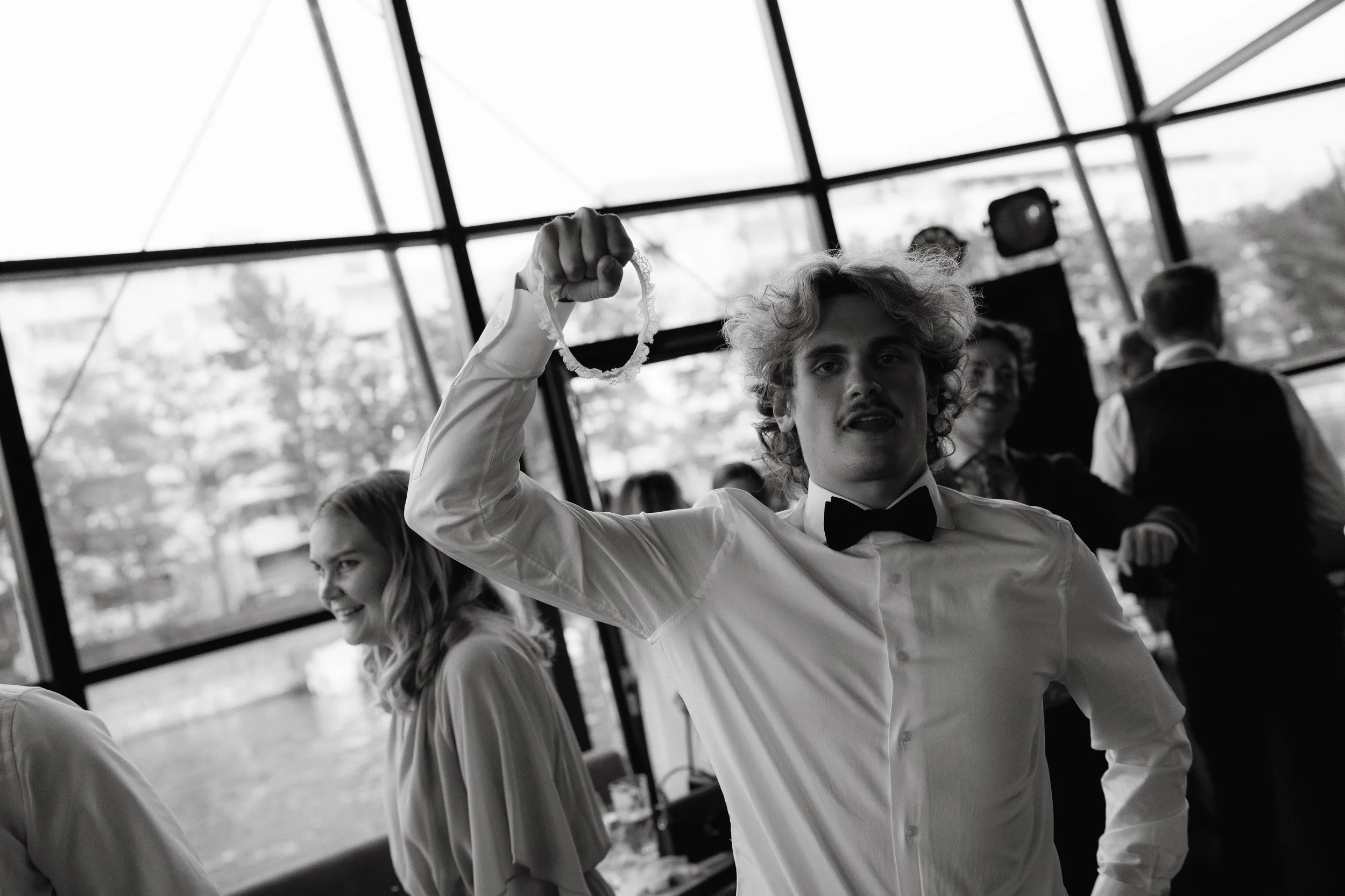 Black and white photo of a man in a formal shirt and bowtie holding a garter, with people in the background in a room with large windows.