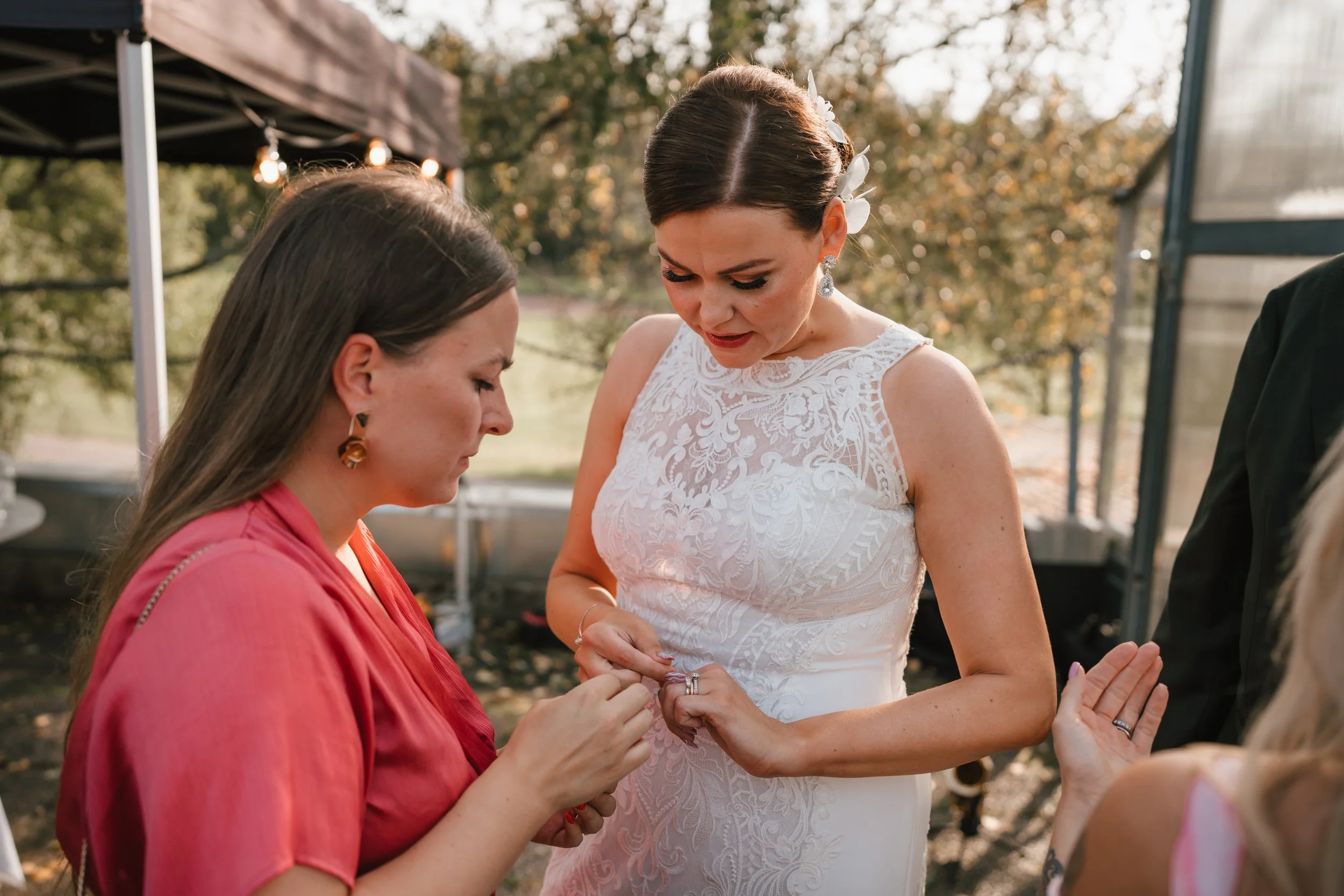 Bride in white lace dress with woman in pink dress outdoors.