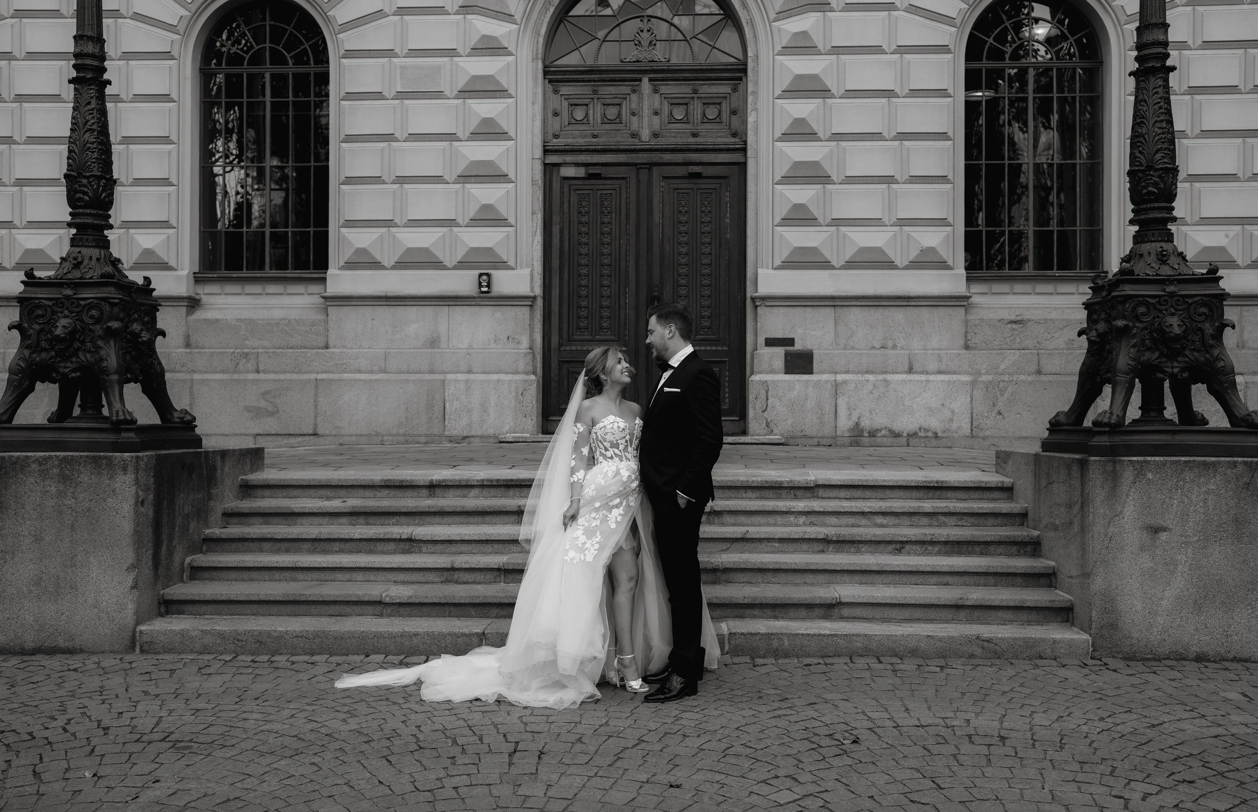 Black and white photo of a bride and groom standing in front of a building with decorative doors and large windows, surrounded by ornate lamp posts.