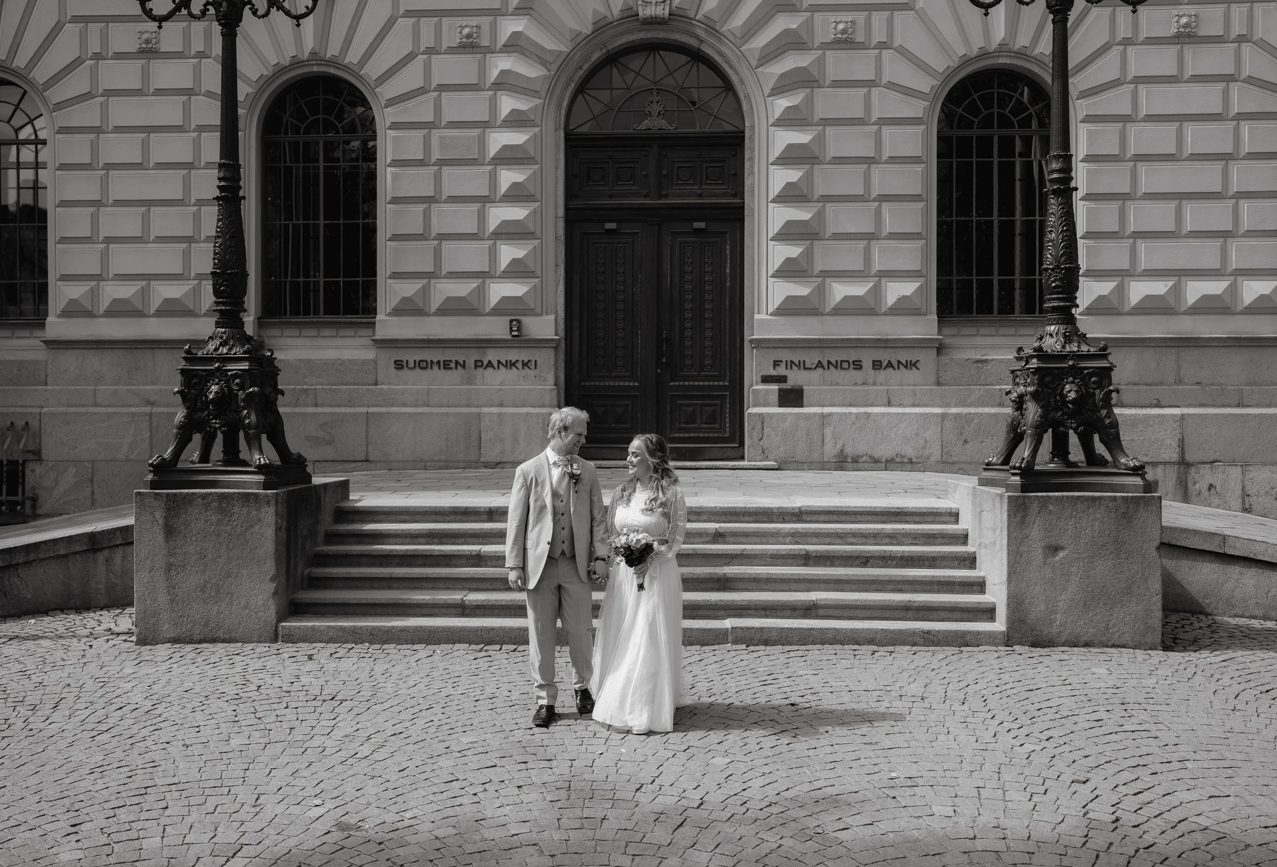 Bride and groom standing outside Finland's Bank, stone steps, ornate street lamps, monochrome image.